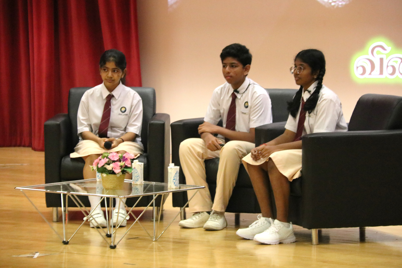 Three students in school uniforms sit on black armchairs on a stage beside a glass table with flowers.