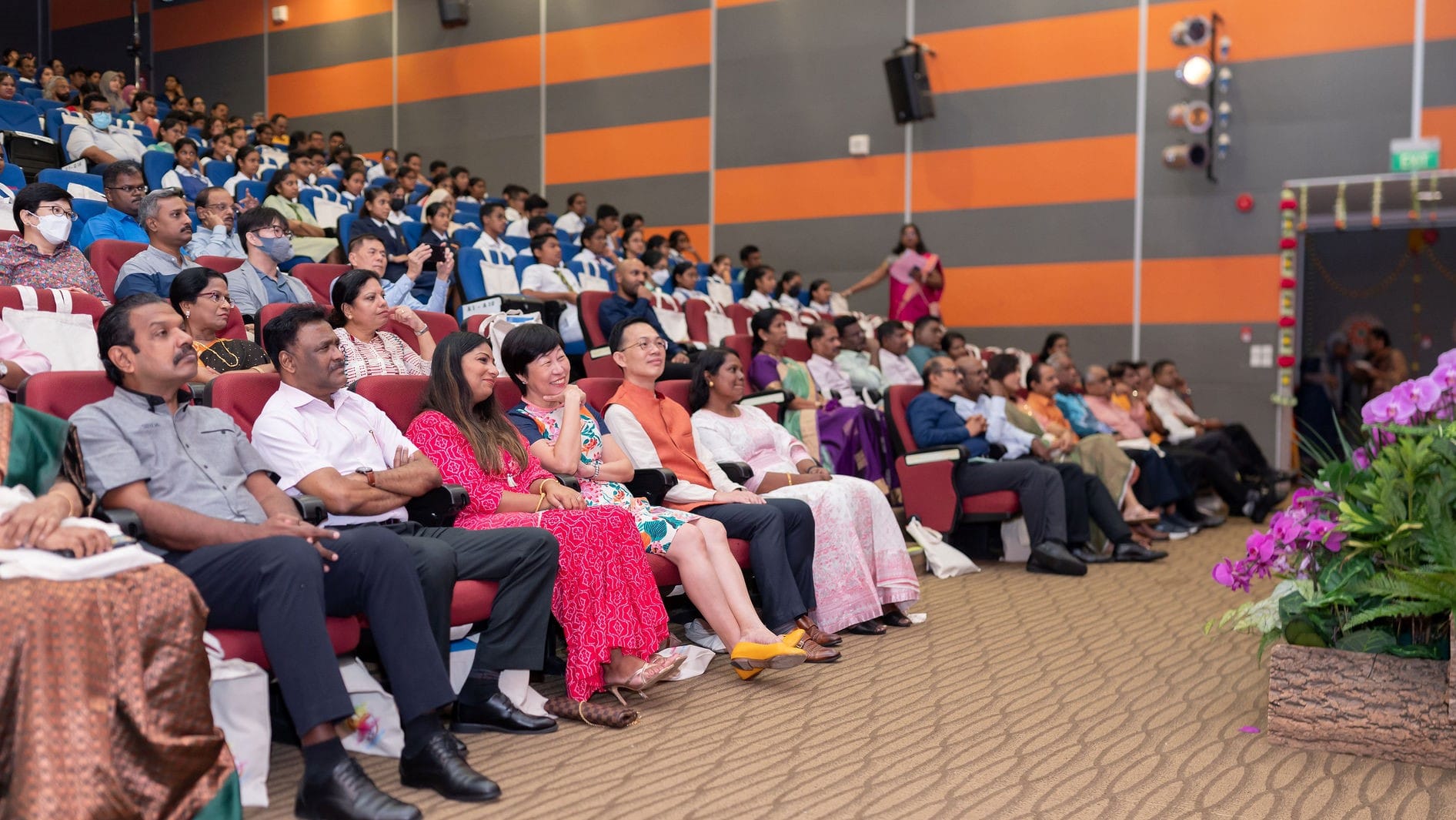 Large auditorium audience seated in rows, with many students in uniform and adults in front seats.