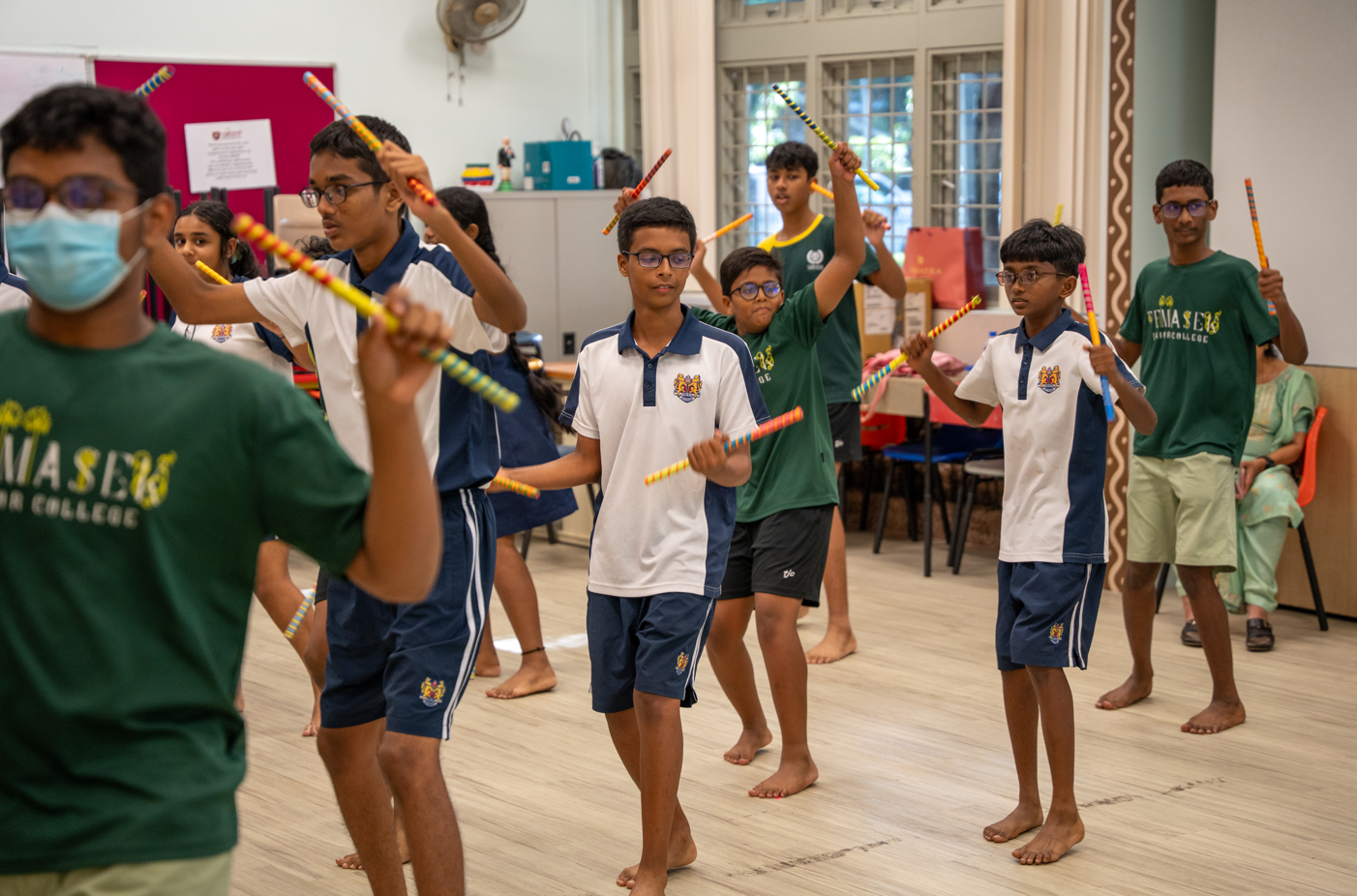 Students practice a stick dance indoors, holding colorful batons; some wear uniforms, one wears a mask.