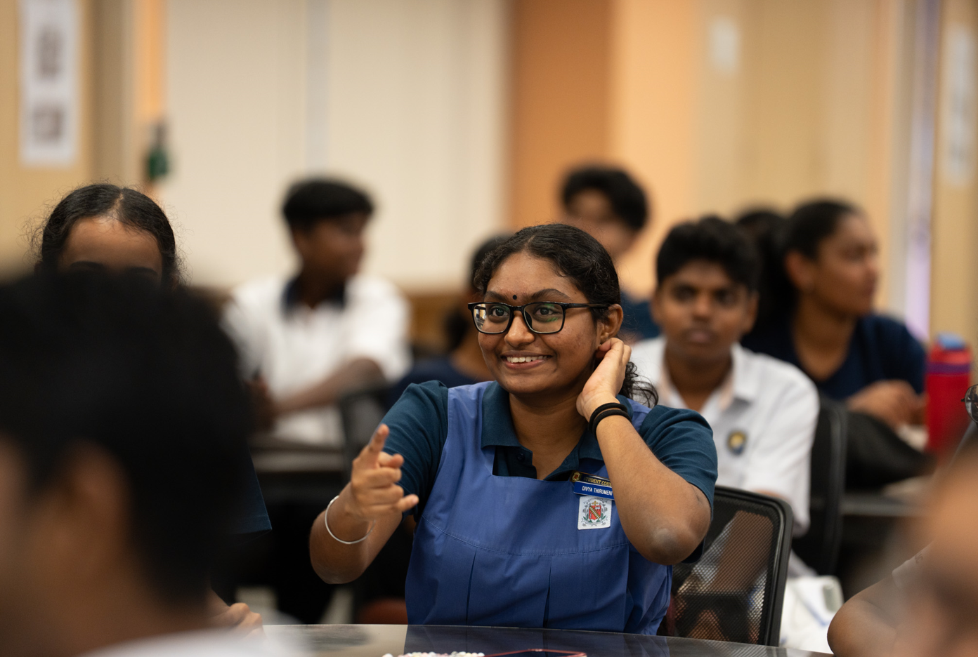 Smiling student in blue uniform and glasses gestures in a classroom, with other students blurred behind.