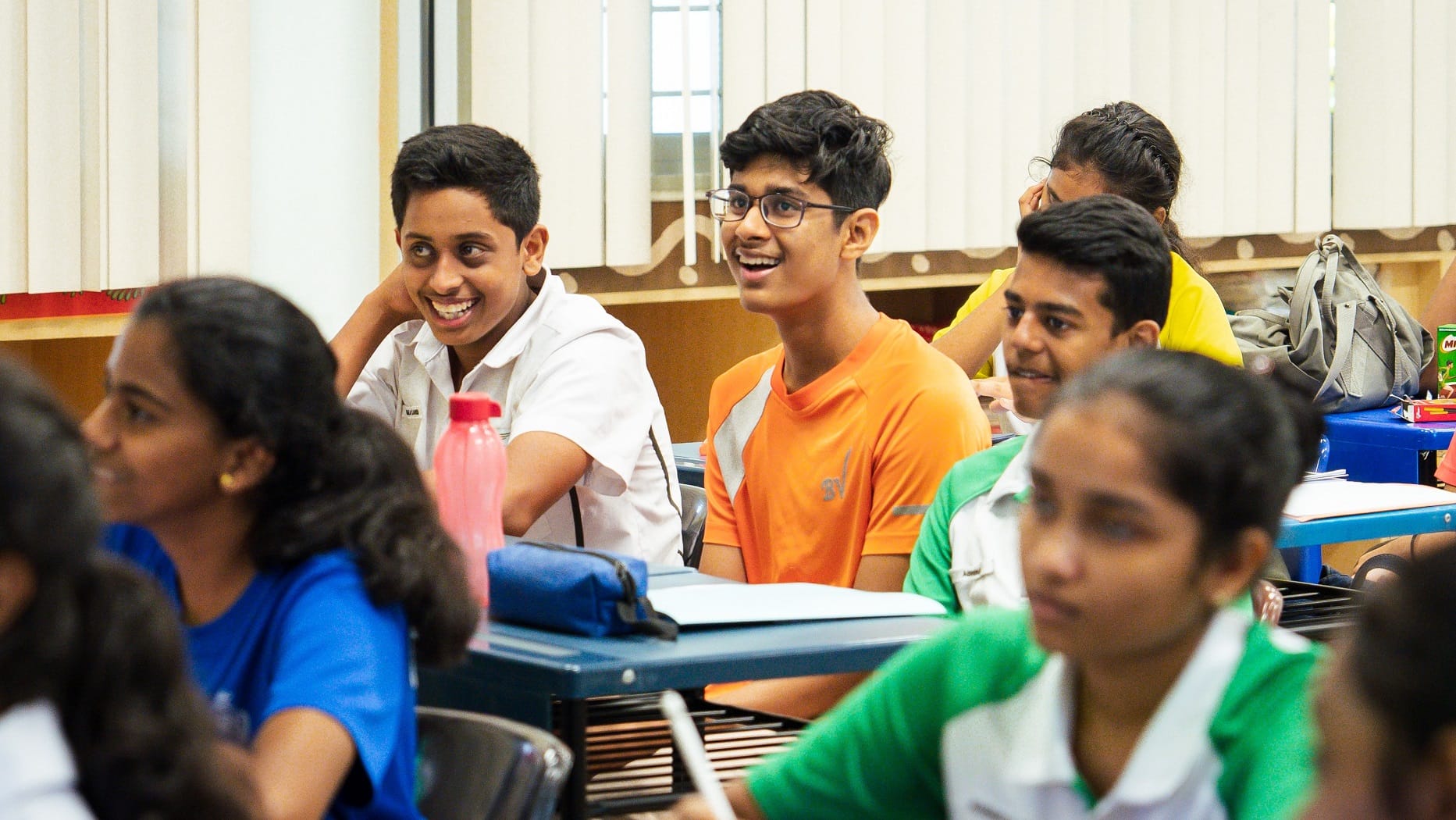 Students seated in a classroom, smiling and looking forward; one wears an orange shirt and glasses.