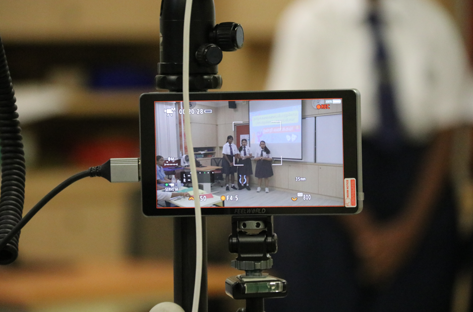 Camera monitor on tripod filming three students presenting in a classroom, with recording overlay on screen.