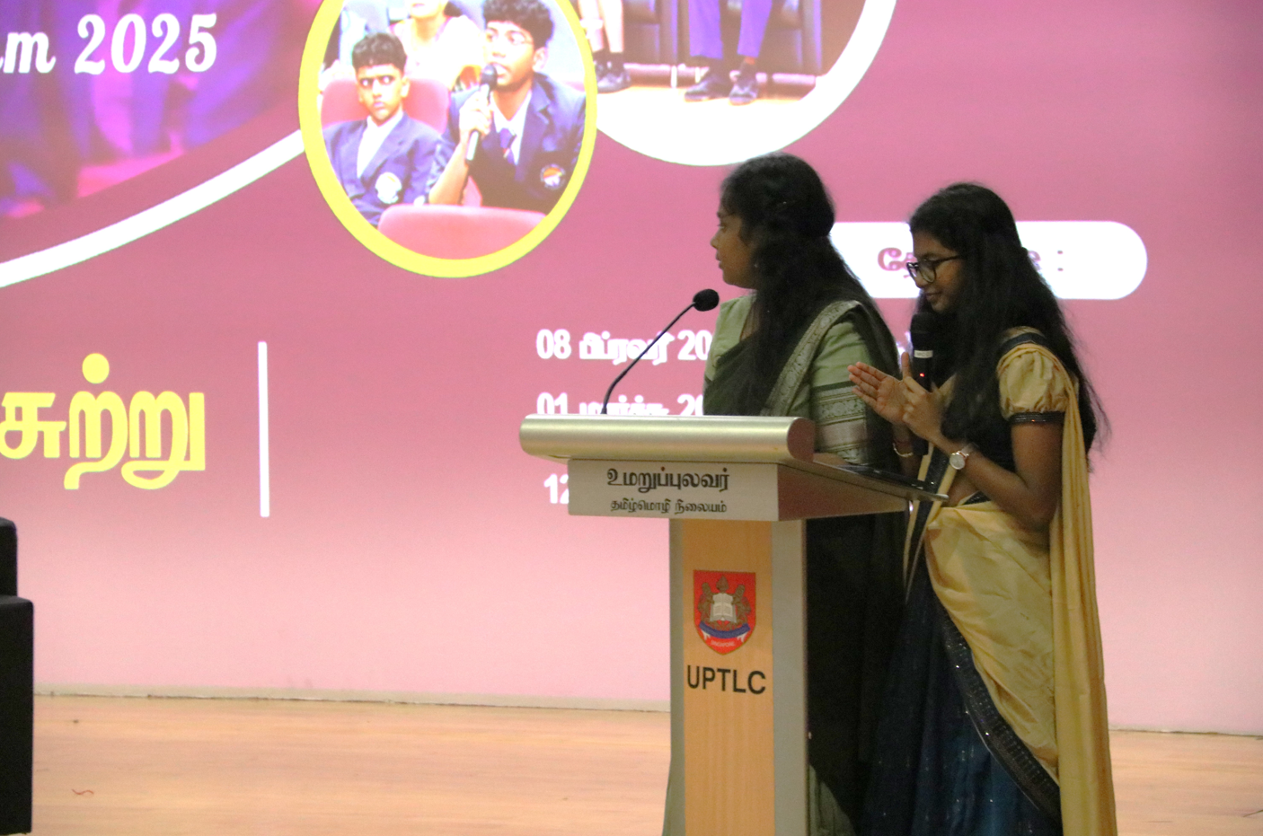 Two women at a podium on stage, one speaking into a microphone, with a large event screen behind.