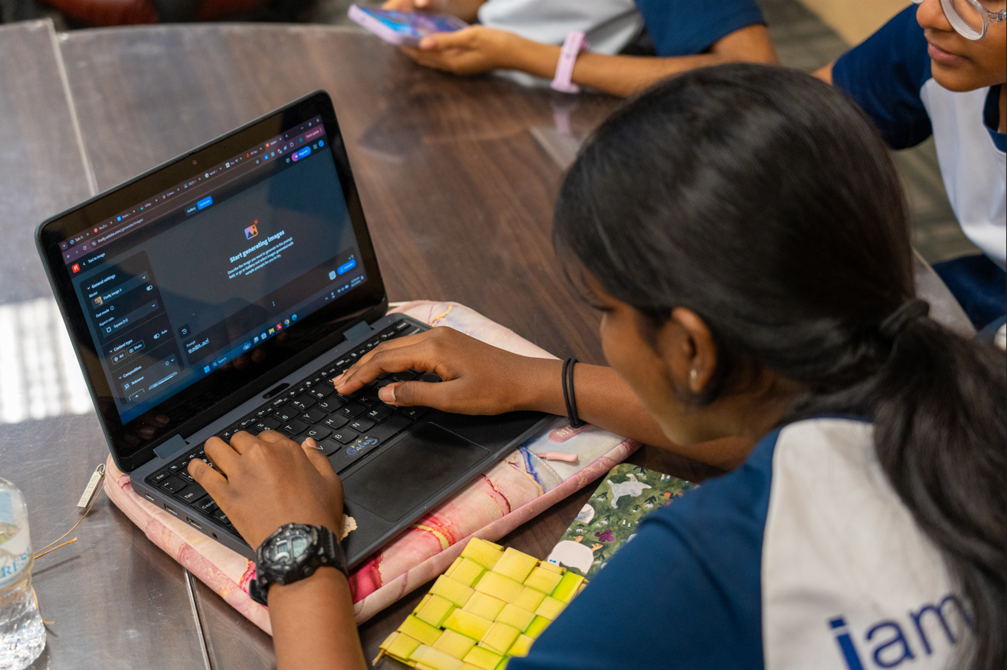 Student with a ponytail types on a laptop at a table while another student looks on.