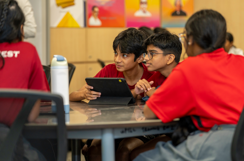 Two boys in red shirts sit at a table, sharing a tablet while classmates sit nearby.