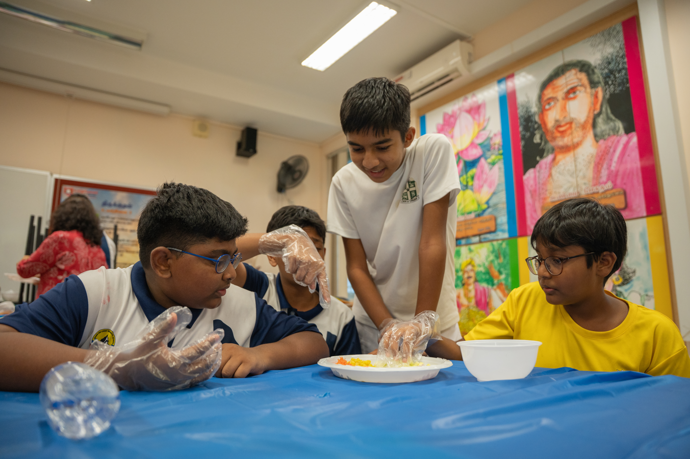 Four boys in a classroom wear plastic gloves and gather around a table with a plate and bowl.