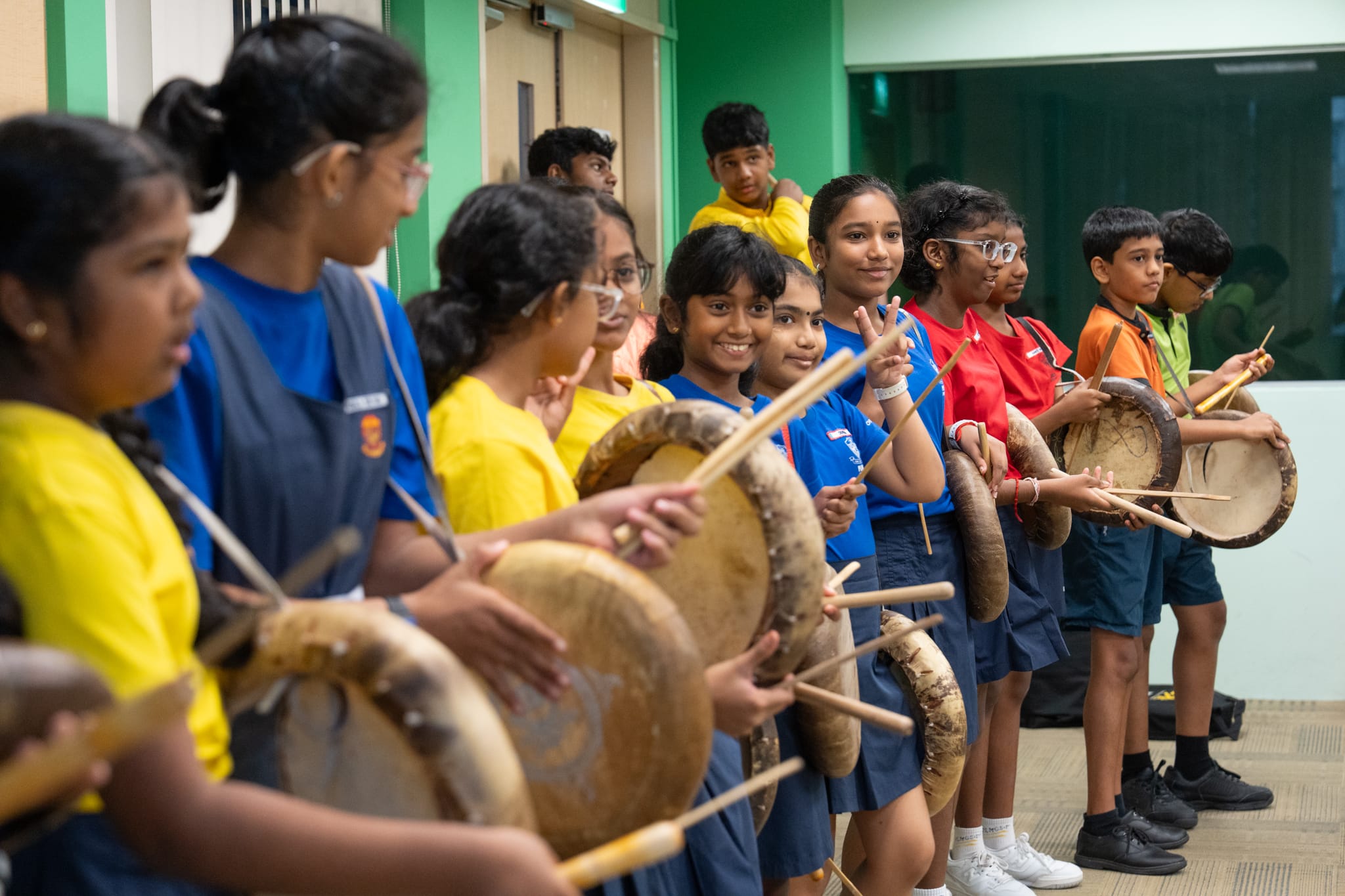 Group of schoolchildren in colorful shirts hold large drums and drumsticks, lined up indoors.