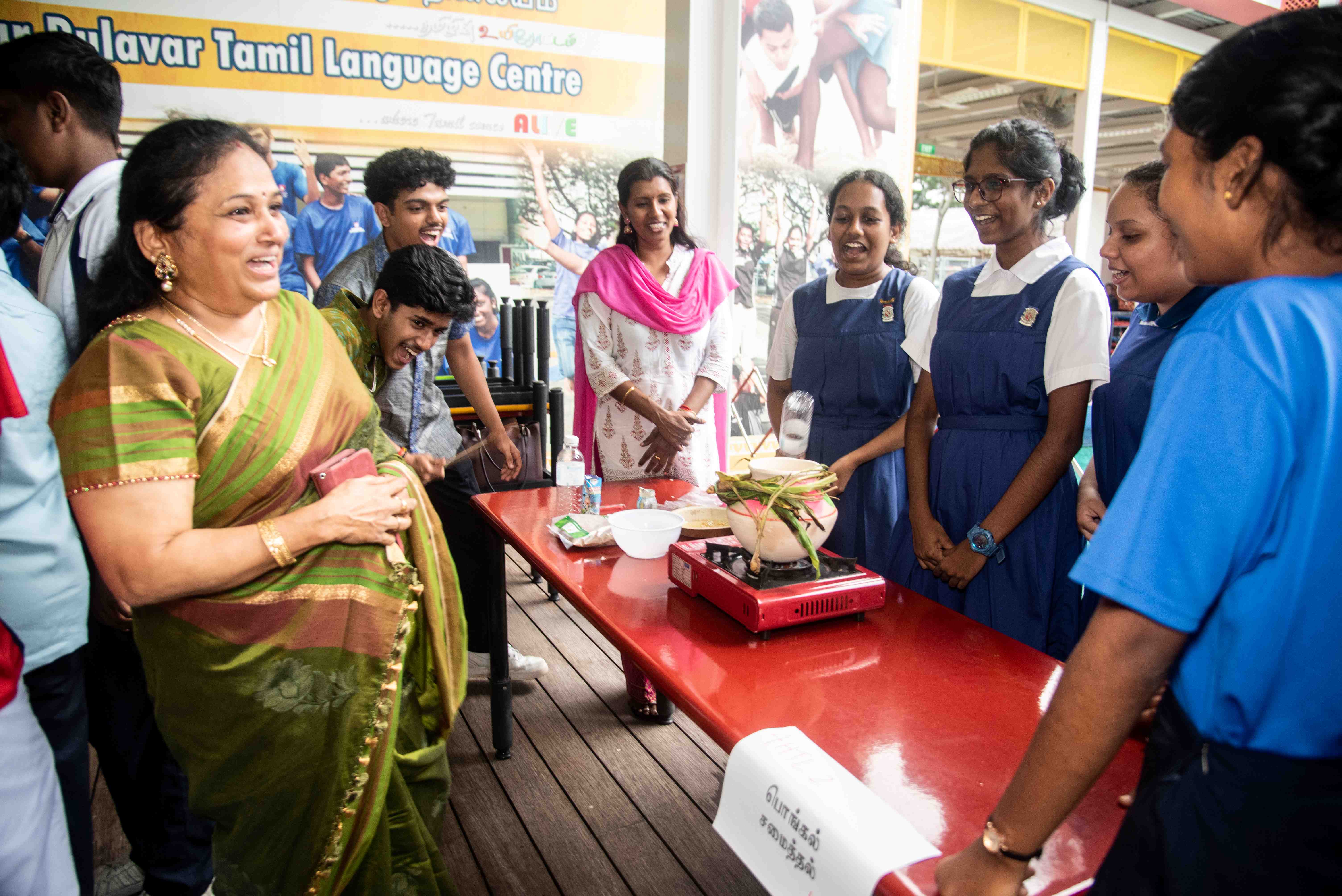 Woman in green sari and students in blue uniforms gathered around a table with a portable stove and pot.