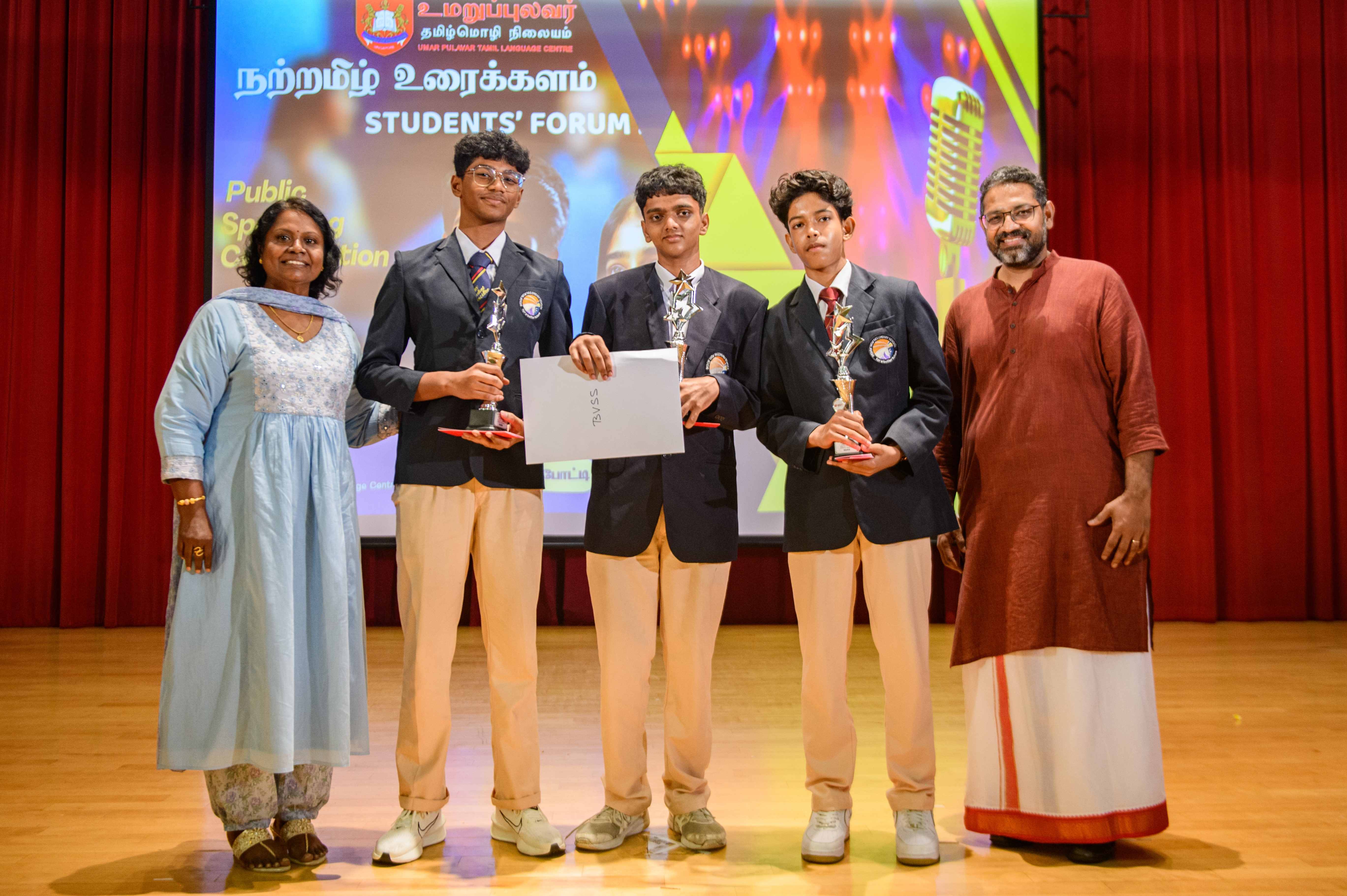 Three schoolboys in blazers hold trophies and a certificate on stage with two adults beside them.