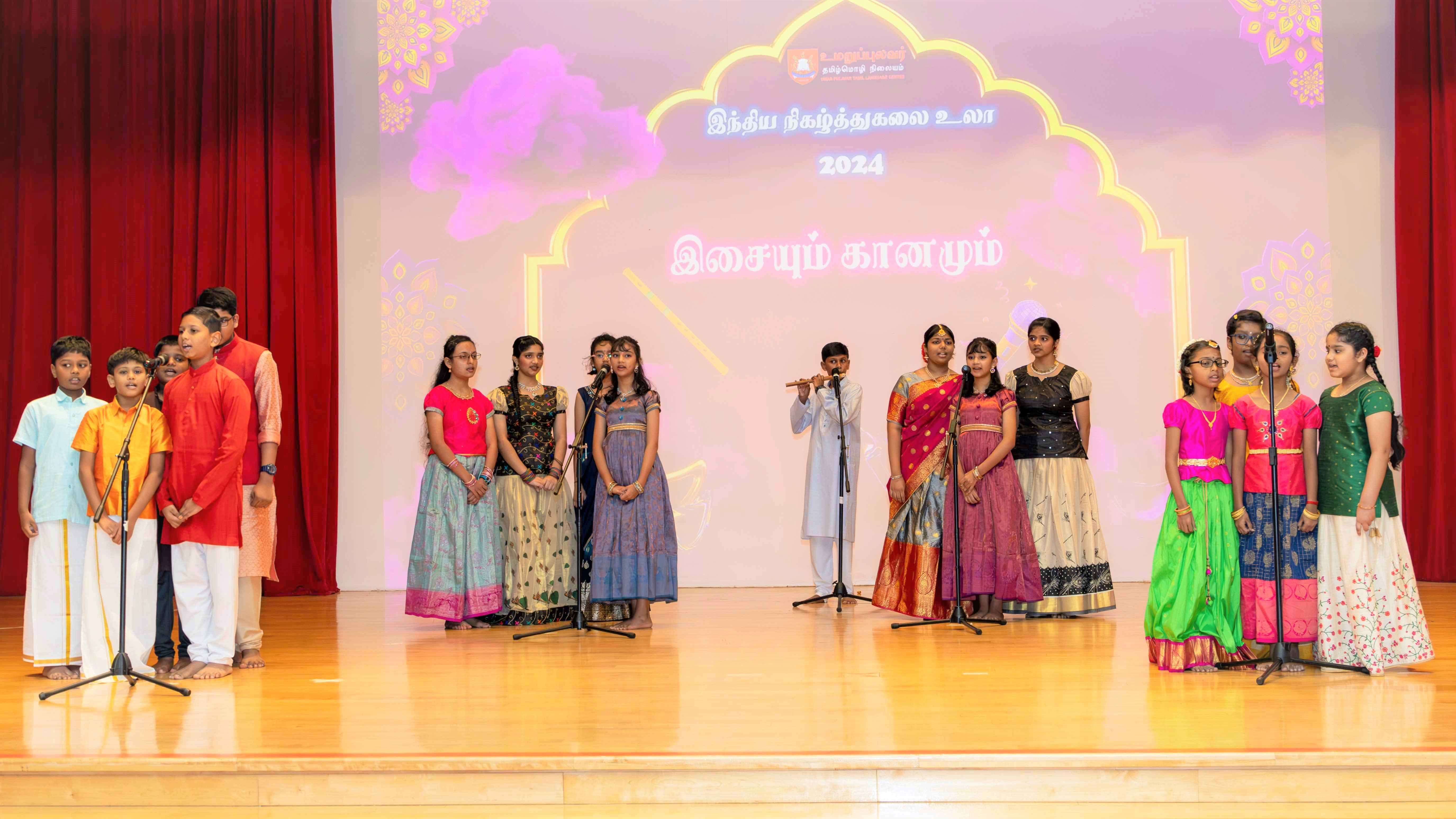 Children in colorful traditional outfits sing on stage at microphones; one boy plays flute under 2024 backdrop.