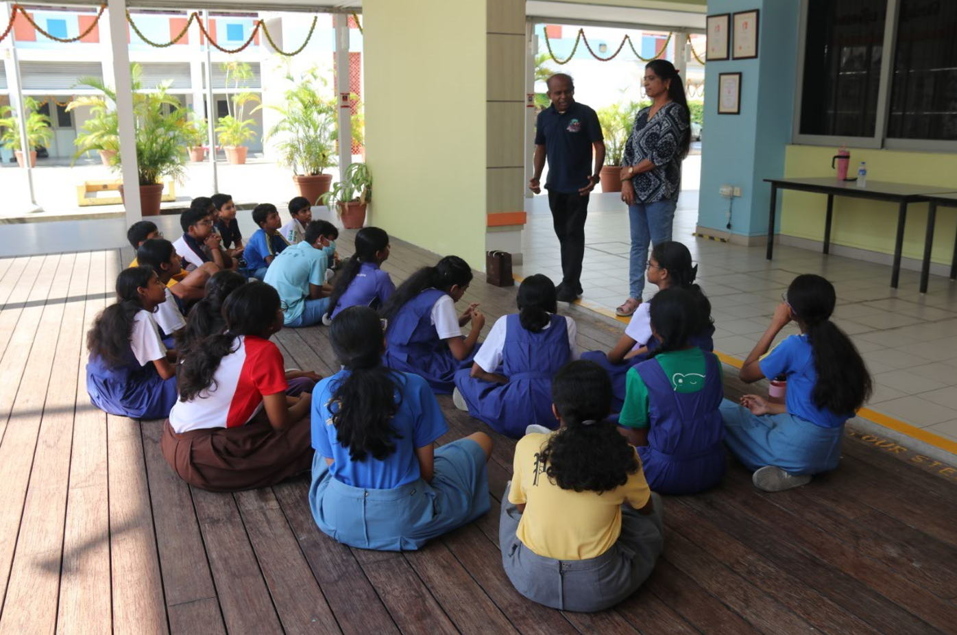 Students seated on a wooden floor listen to two adults speaking in a school corridor with potted plants.