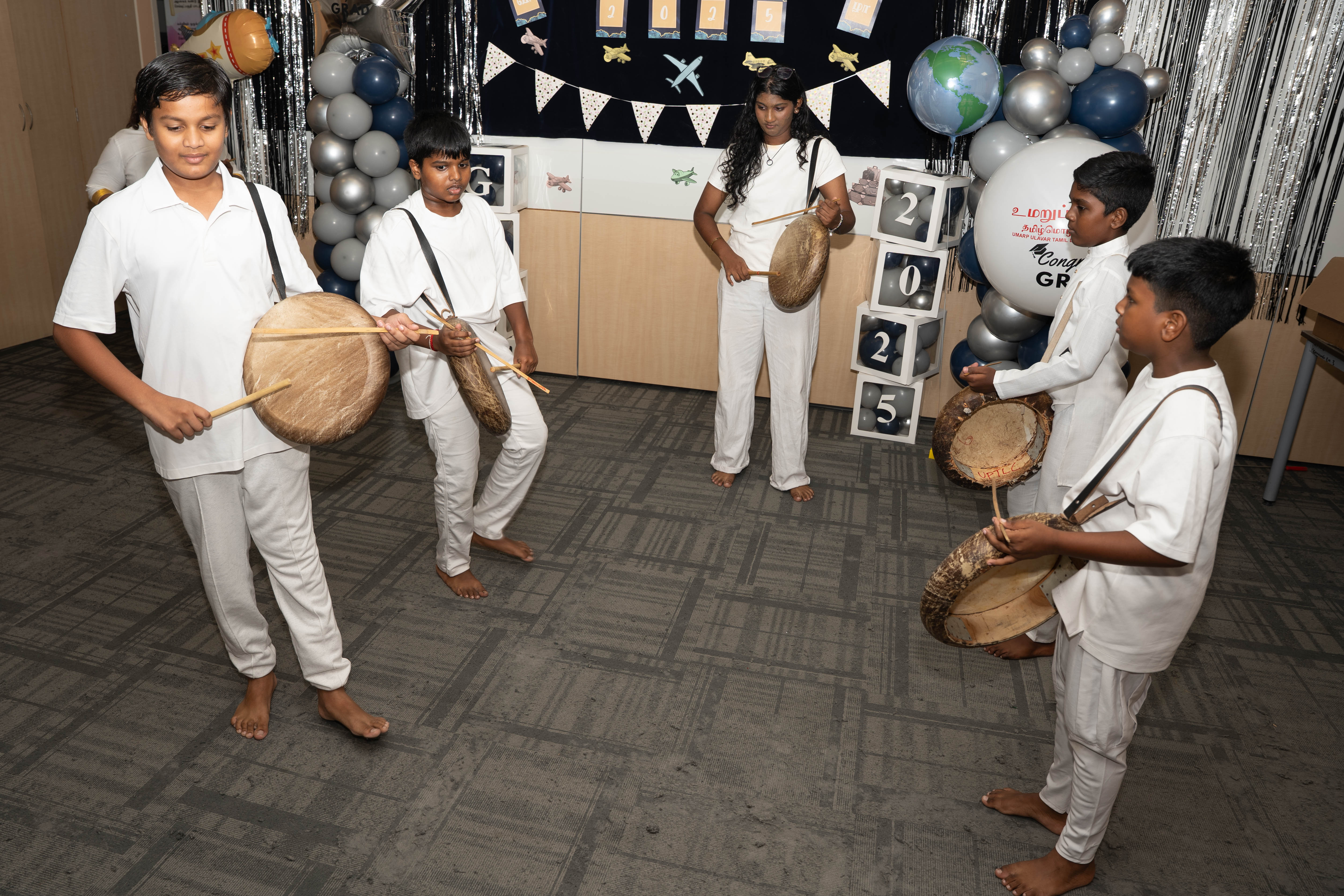 Five kids in white outfits play frame drums with sticks at a decorated party backdrop with balloons.