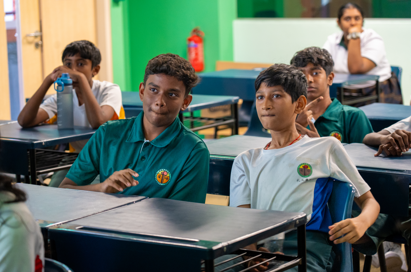 Students in uniforms sit at desks in a classroom, looking forward; green wall and fire extinguisher behind.