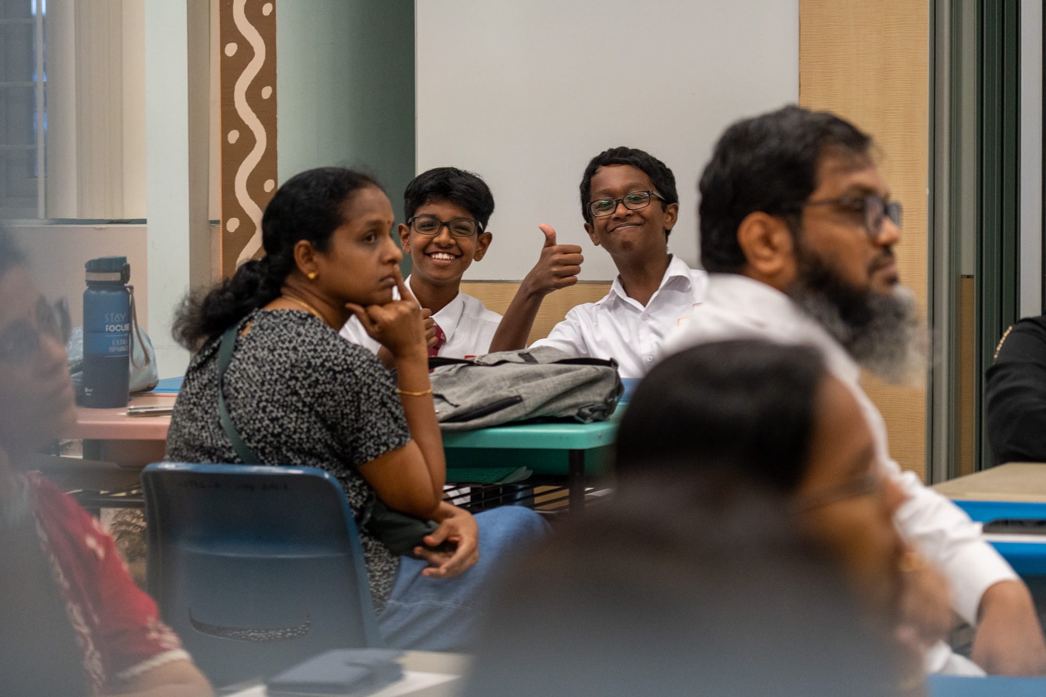 Two schoolboys in white shirts smile at a table; one gives a thumbs-up as adults sit in foreground.