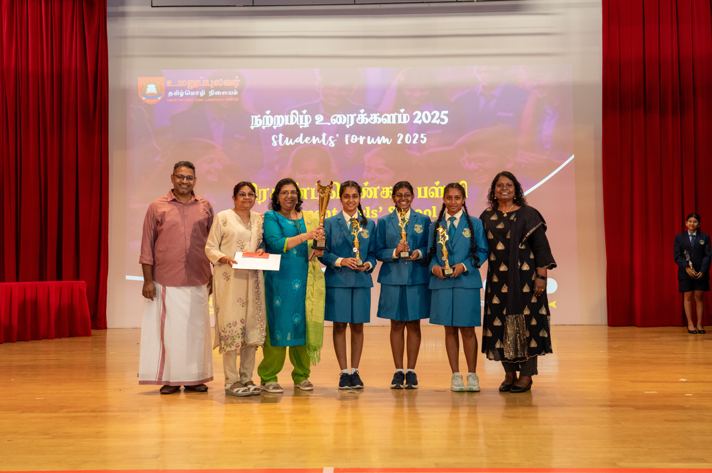 Group on stage; three girls in blue uniforms hold trophies in front of a screen reading Students Forum 2025
