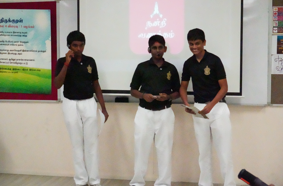 Three students in black shirts and white pants stand before a projector screen, one holding a mic.