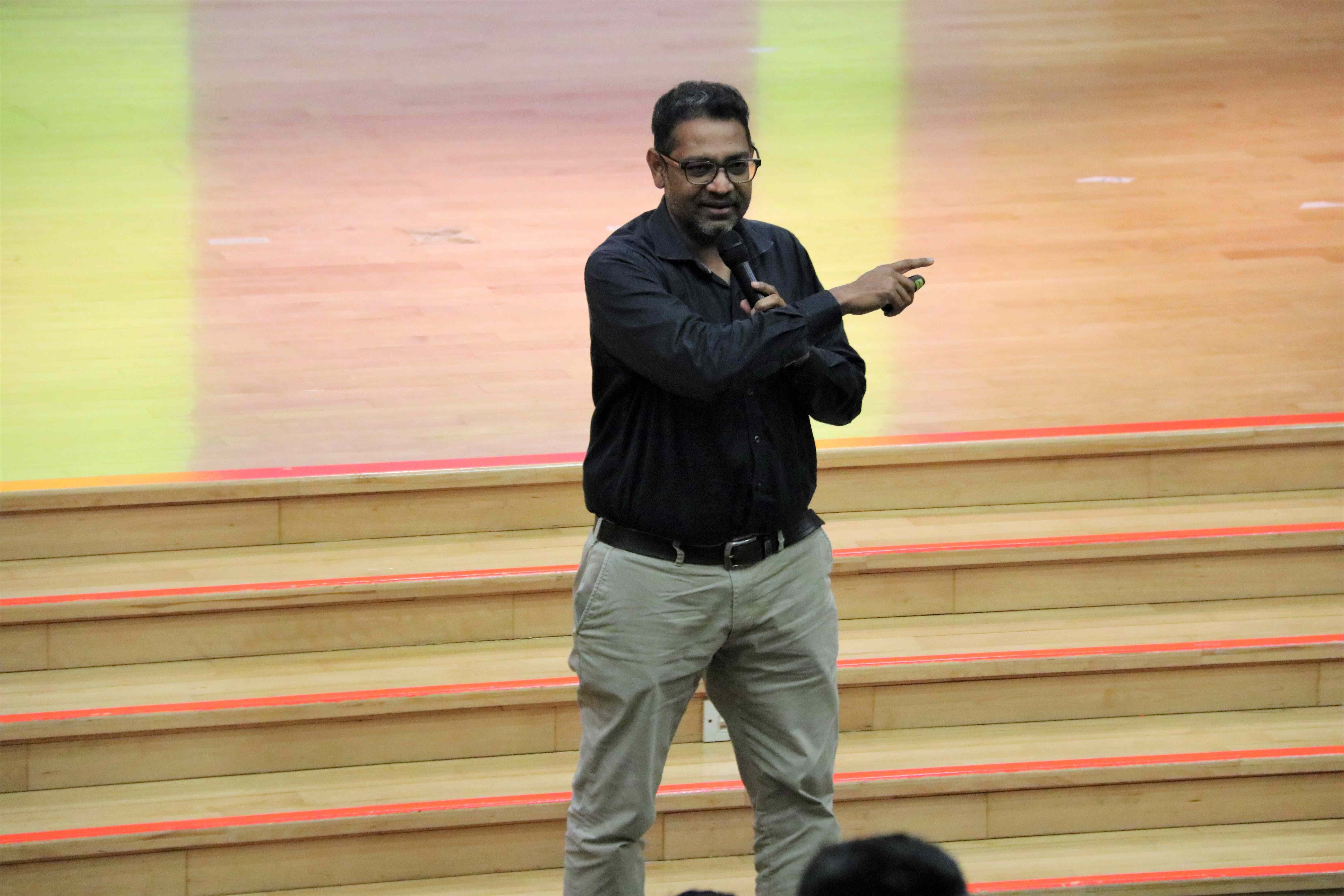 Man in glasses holding a microphone and pointing right while standing on steps in an auditorium.