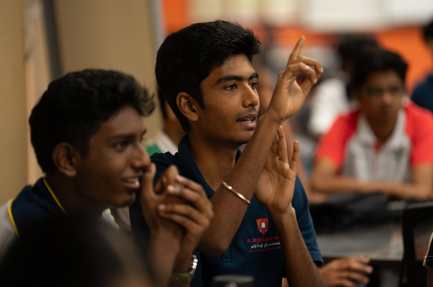 Young man in a navy shirt raises a finger while speaking, with other students blurred in background.