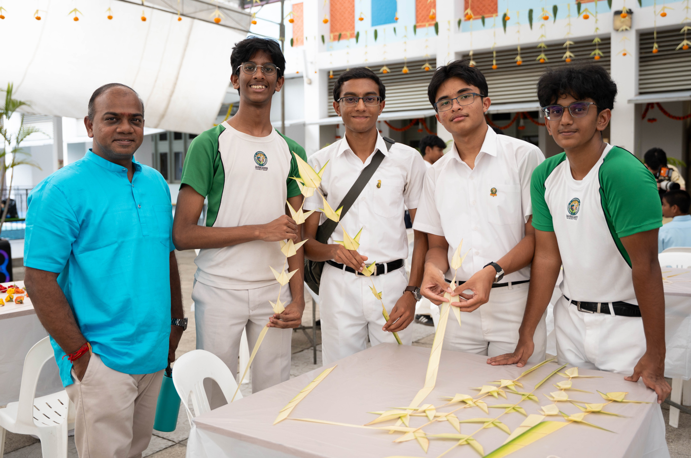 Man in blue shirt stands with four students in white uniforms, holding palm leaf crafts at a table.