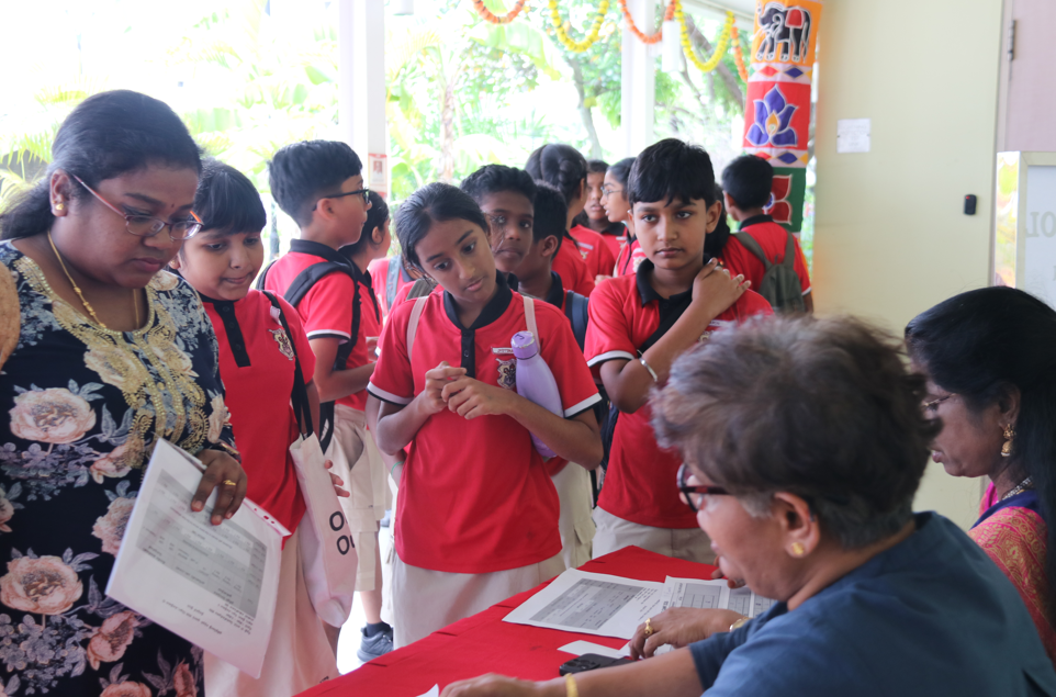 Students in red uniforms line up at a table as adults check papers at a school event.