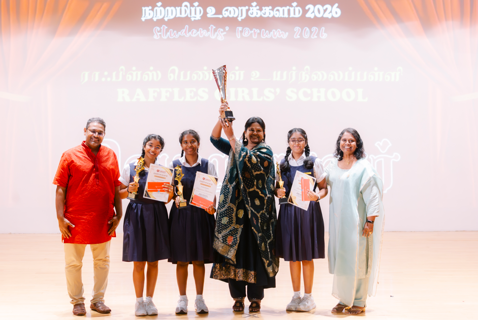 Six people on stage; three schoolgirls hold trophies and certificates, woman in center raises a cup.