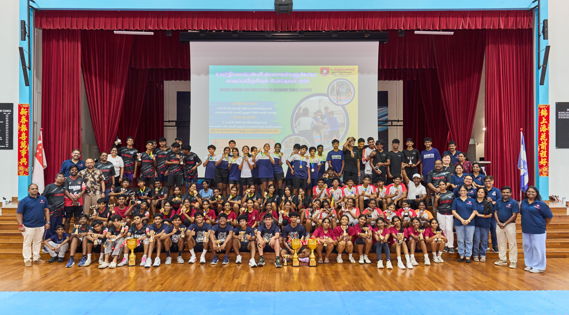 Large group of students and adults posing on a school stage holding trophies and medals at a kabaddi competition.