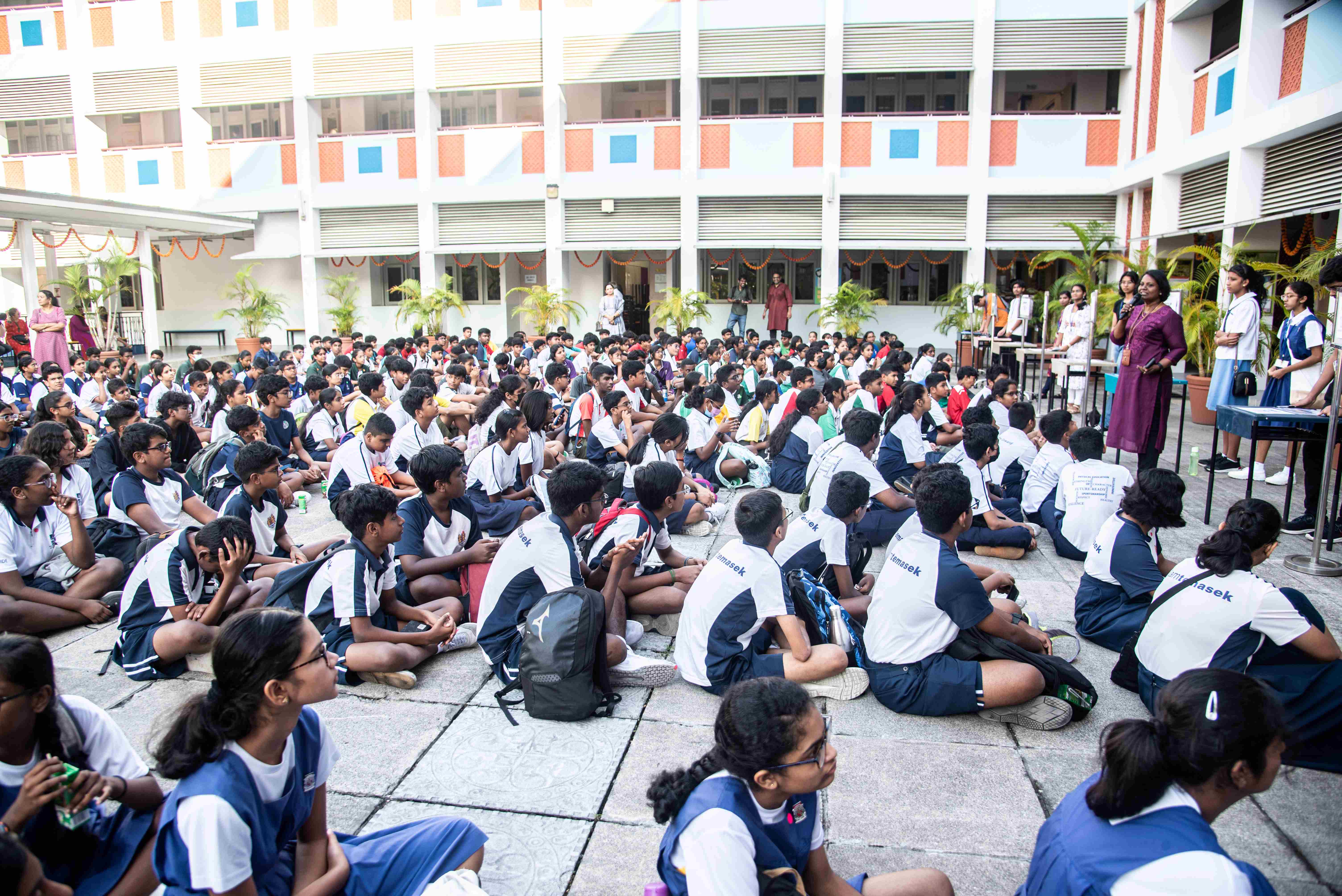 Large group of students in uniforms sit in a courtyard facing teachers near a microphone stand.