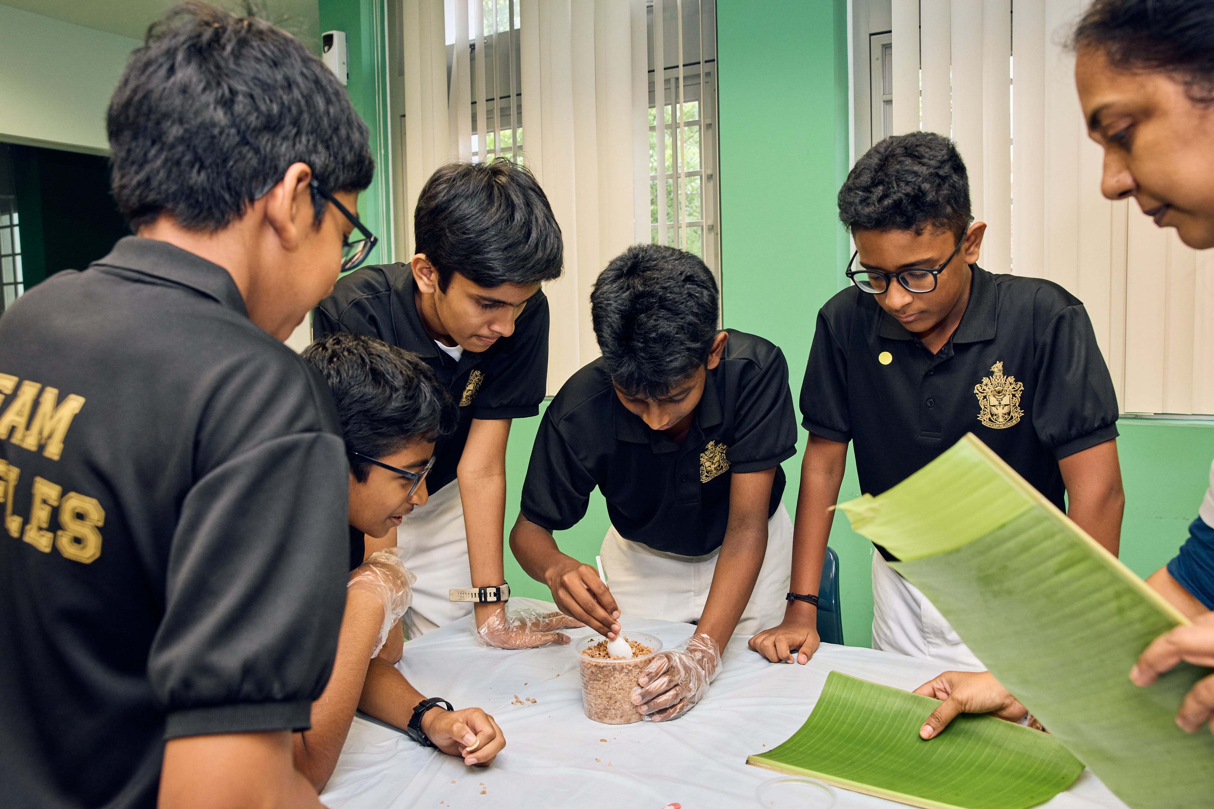 Five students in black shirts gather around a table as one mixes food in a clear container.
