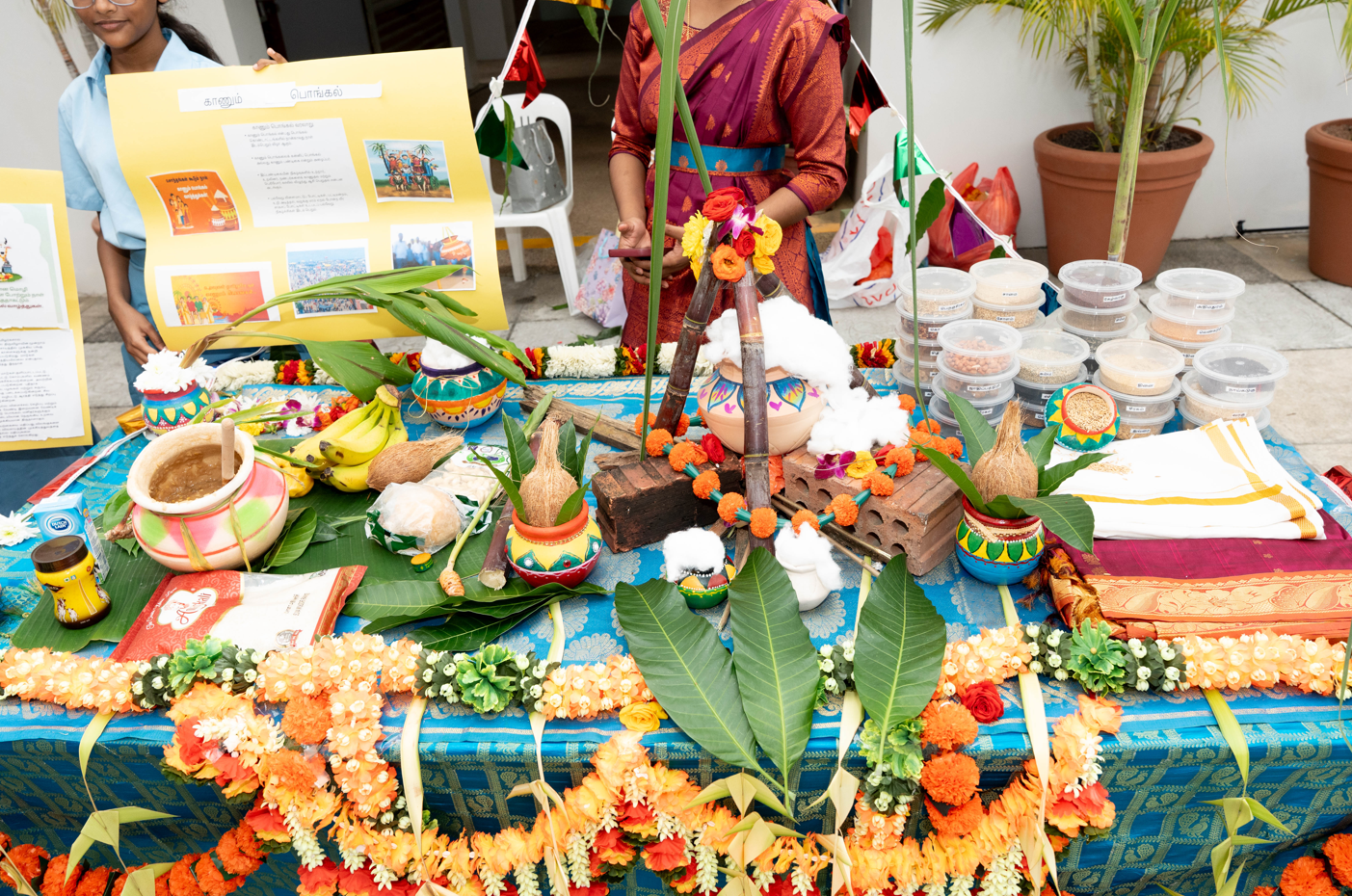 Decorated table with clay pots, coconuts, bananas, flowers, and bowls of food; people stand behind.