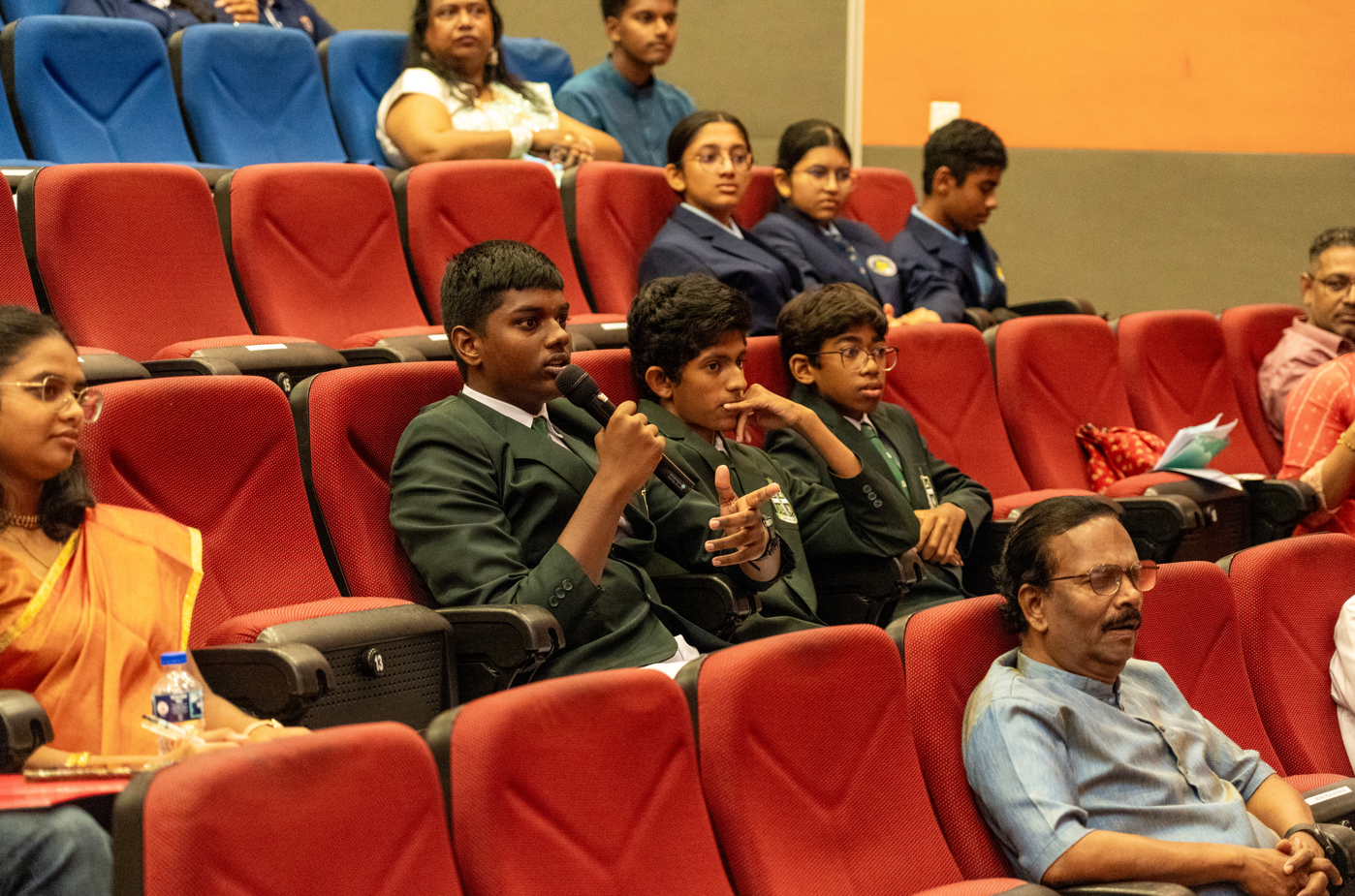 Student in green blazer speaks into a microphone while seated among red auditorium chairs with others listening.