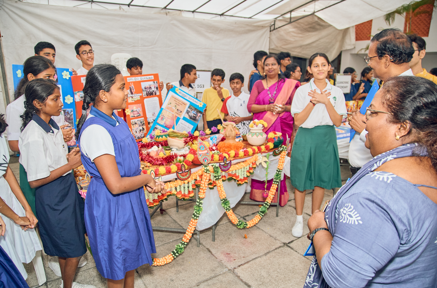 Students in uniforms present a flower-decorated display table to teachers under a canopy, with posters behind.
