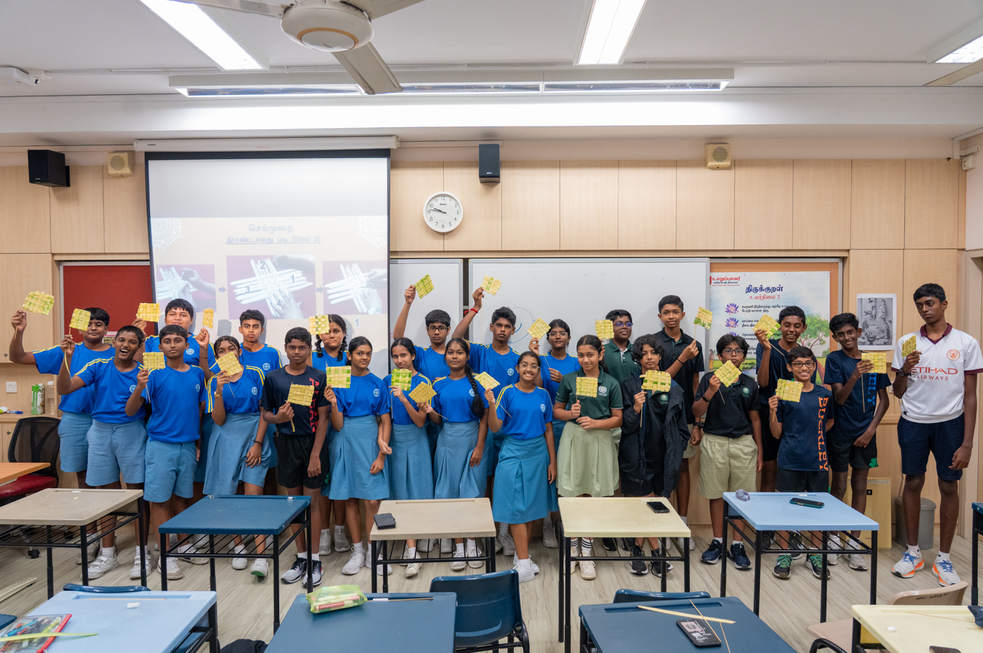 Group of students in a classroom posing and holding up small yellow woven crafts in front of a screen.