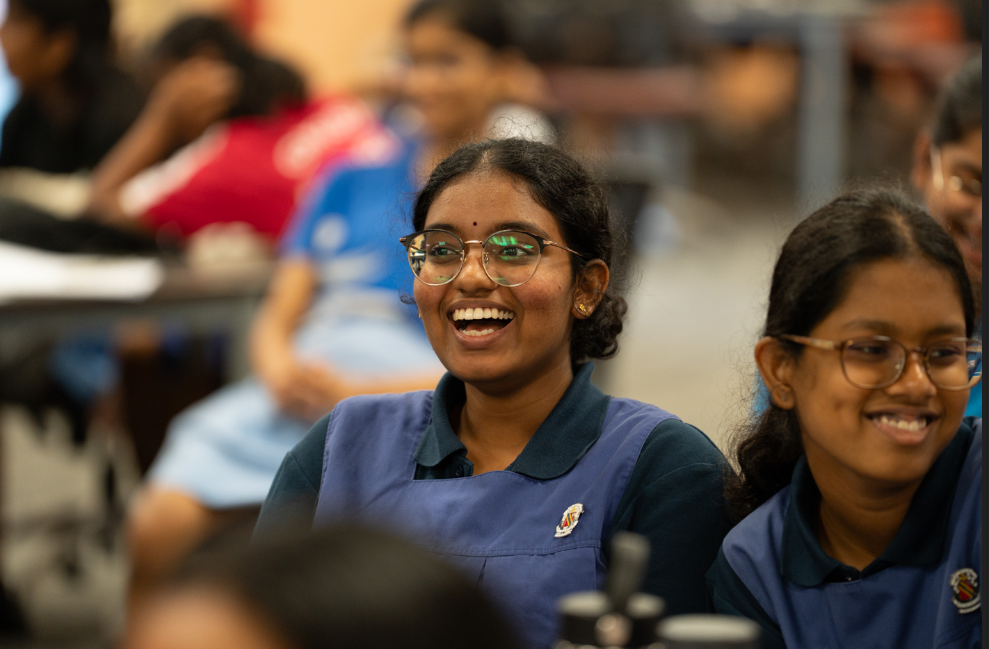 Smiling student in glasses and blue uniform sits in a classroom, with other students blurred behind.