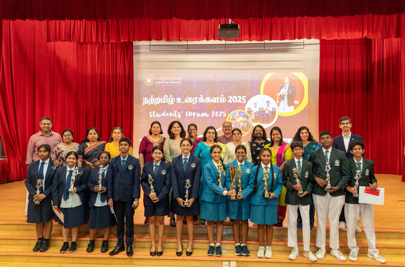 Students in uniforms hold trophies on a stage at Students' Forum 2025, with adults standing behind them.