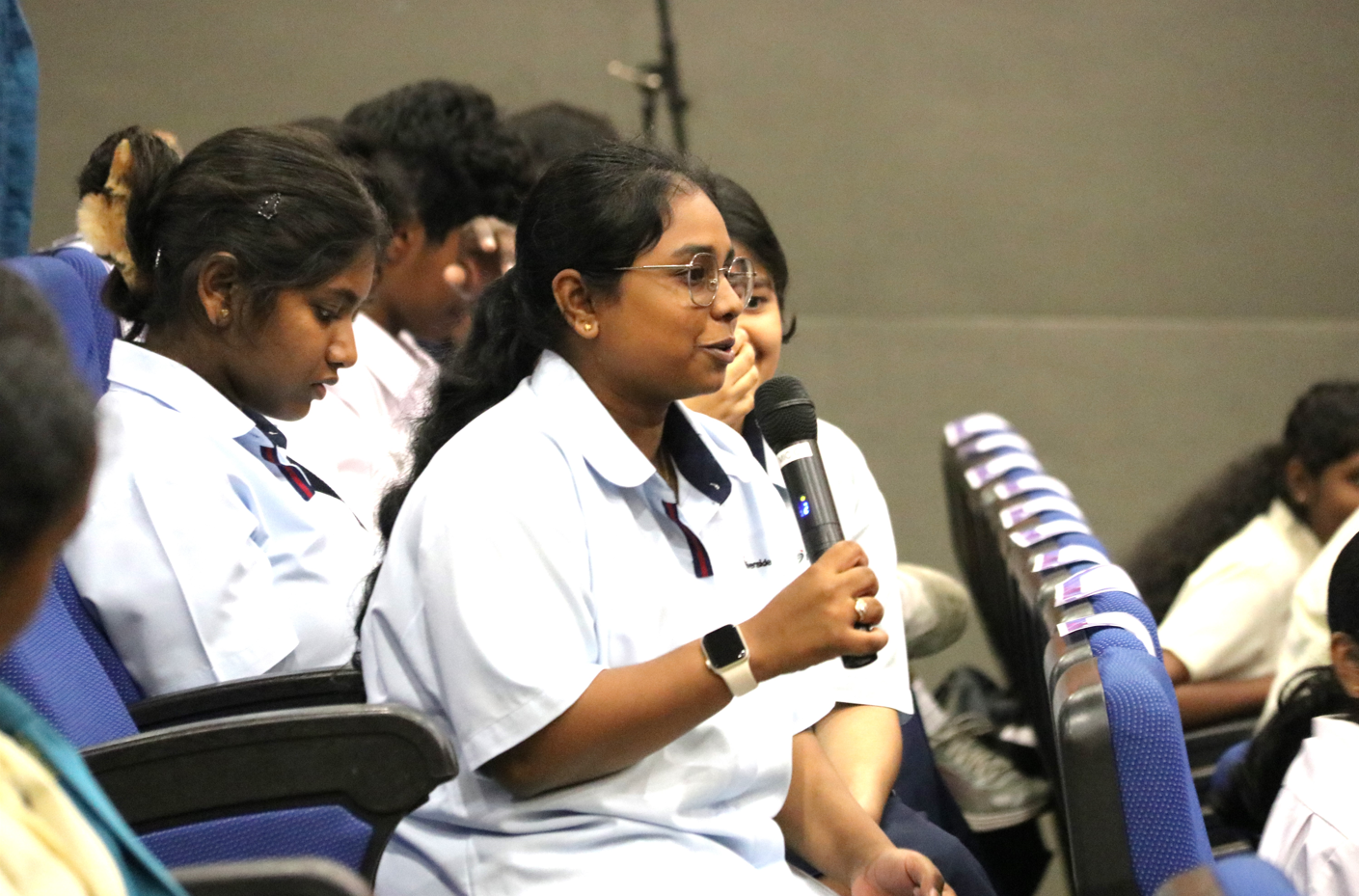 Student in a white uniform and glasses speaks into a microphone while seated in an auditorium.