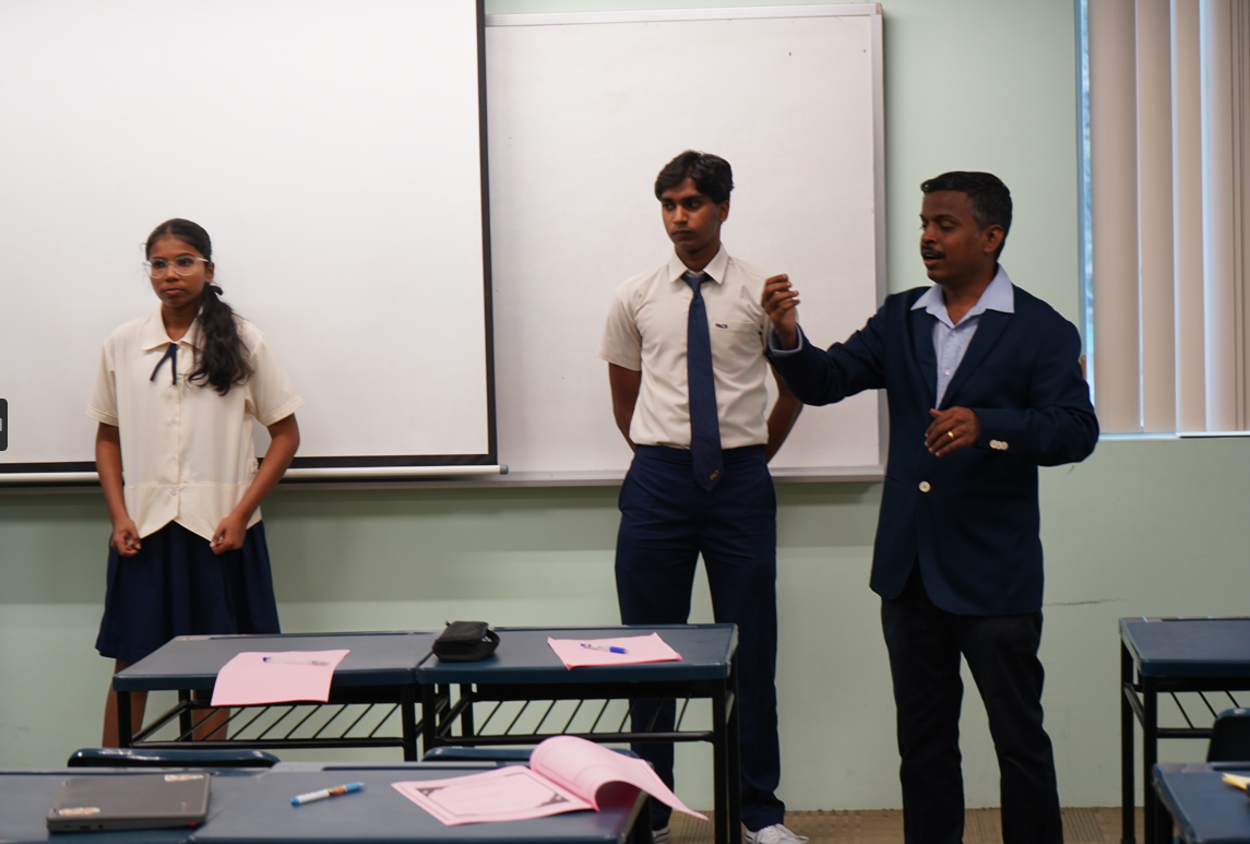 Man in suit gestures while speaking beside two students in uniform at front of classroom near whiteboard.