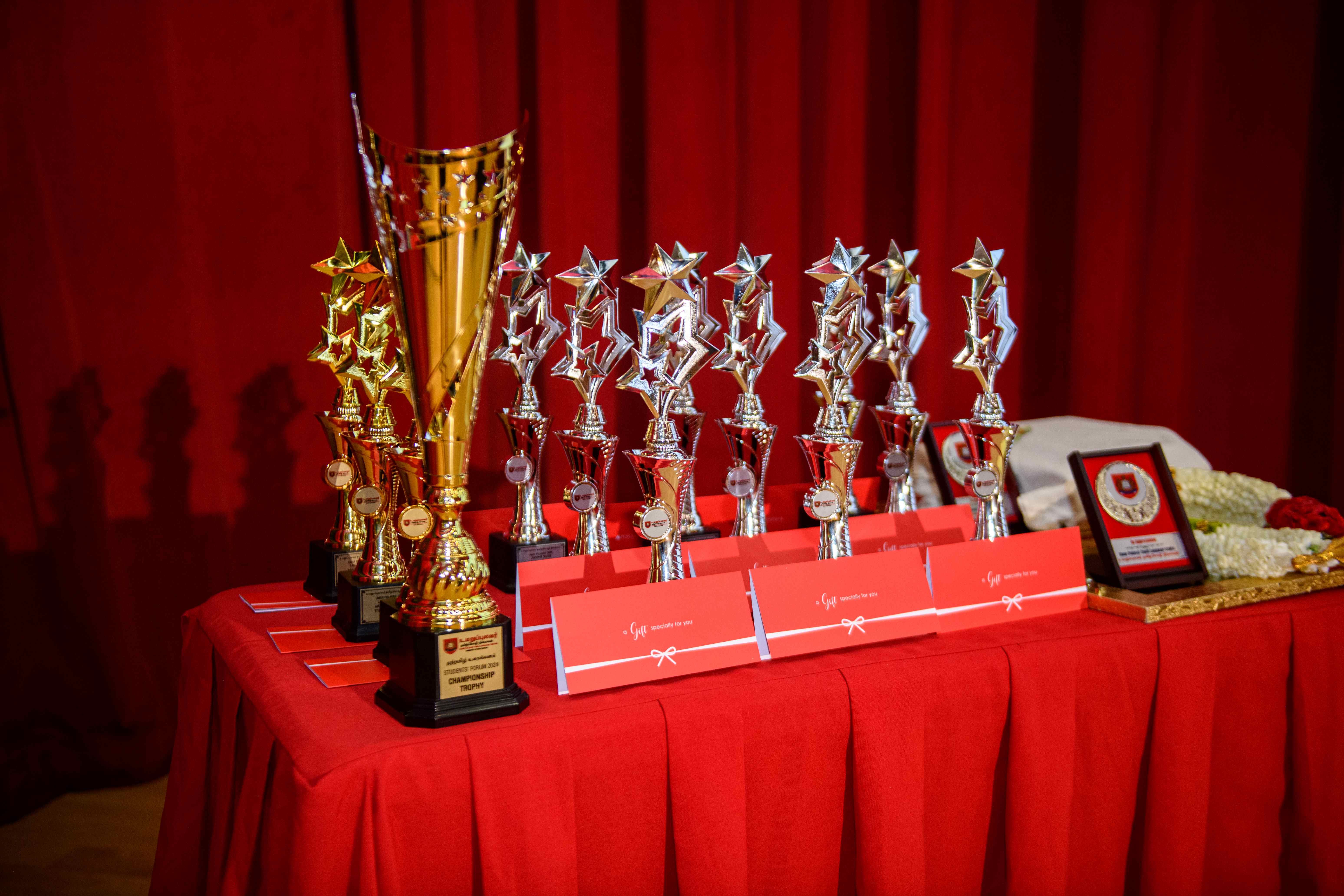 Gold and silver star trophies and red envelopes on a red-covered table in front of red curtains.