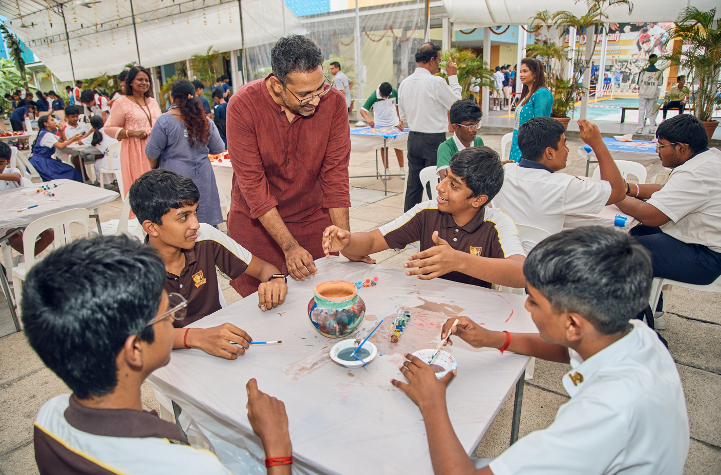 Teacher in red kurta leans over students painting a clay pot at a table in a busy outdoor school fair.