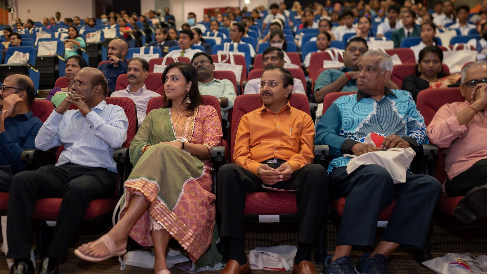 Front row of seated adults in an auditorium, with many attendees in blue and red seats behind.