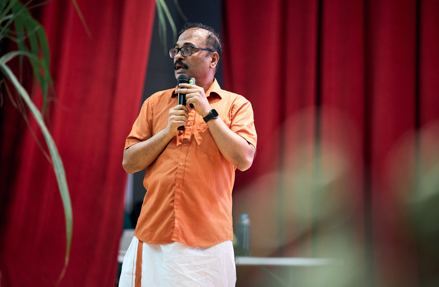 Man in orange shirt and white wrap holds a microphone on stage before red curtains, looking sideways.