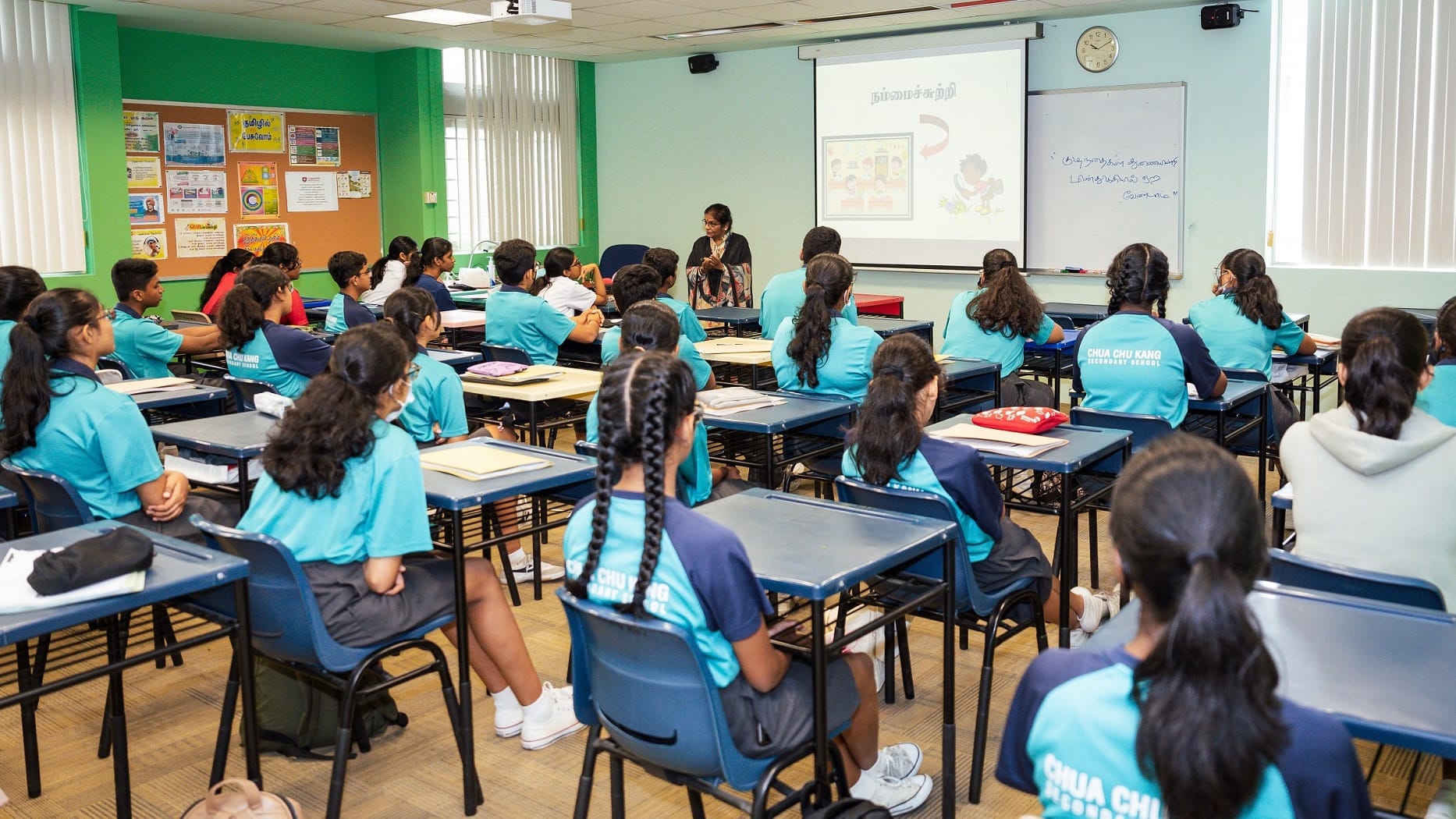 Teacher stands at front as students in blue uniforms sit at desks facing a projector screen and whiteboard.
