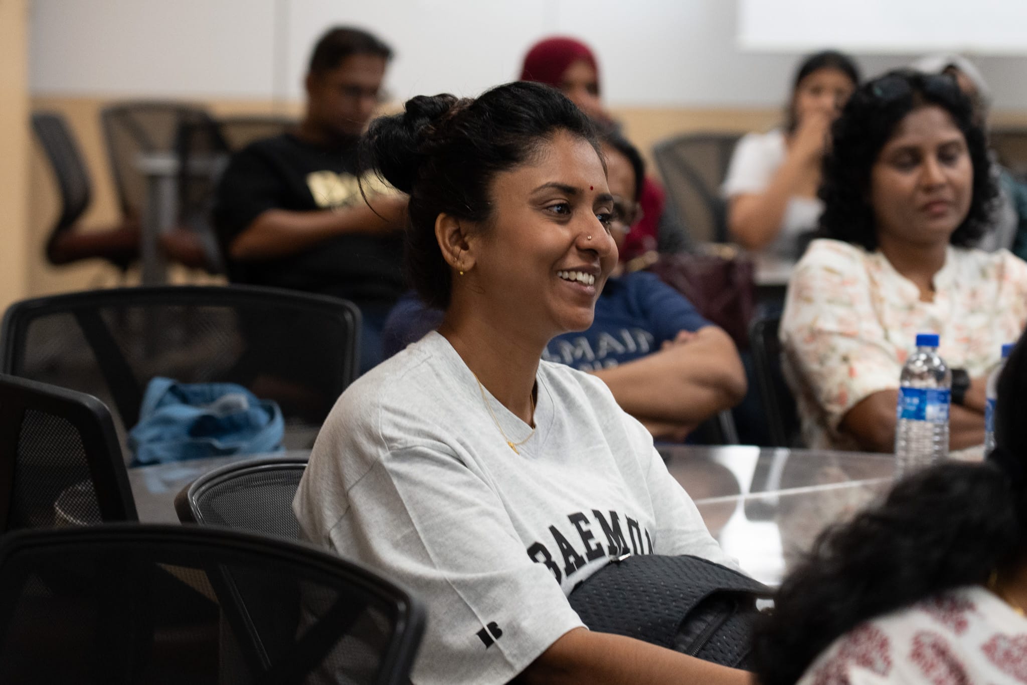 Smiling woman with hair in a bun sits at a table in a classroom, with others seated behind her.