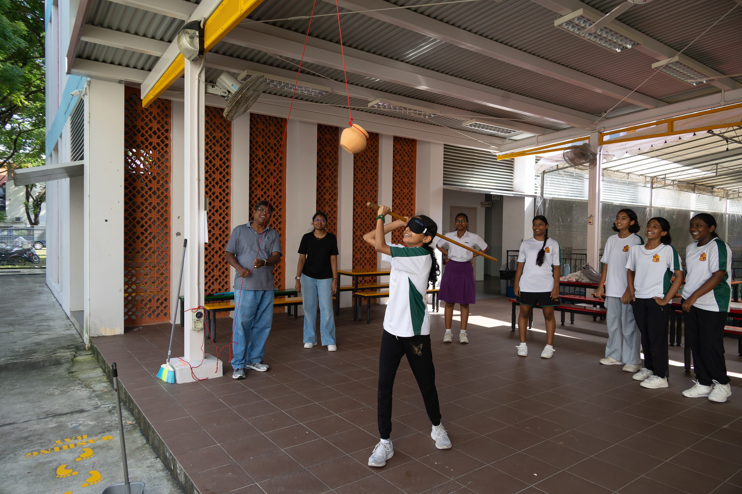 Student wearing a blindfold swings a stick at a hanging pot as classmates watch under a covered school area.