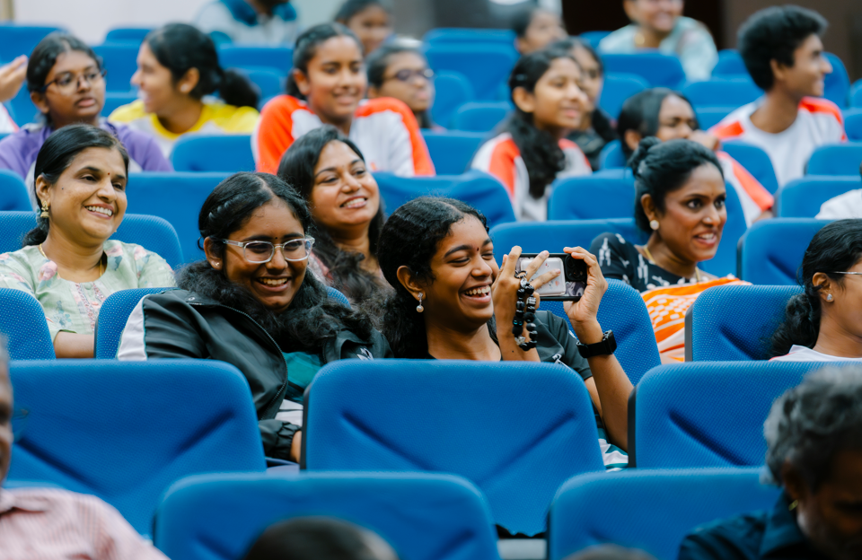 Audience seated in blue auditorium chairs, smiling and laughing; one woman holds up a phone to record.