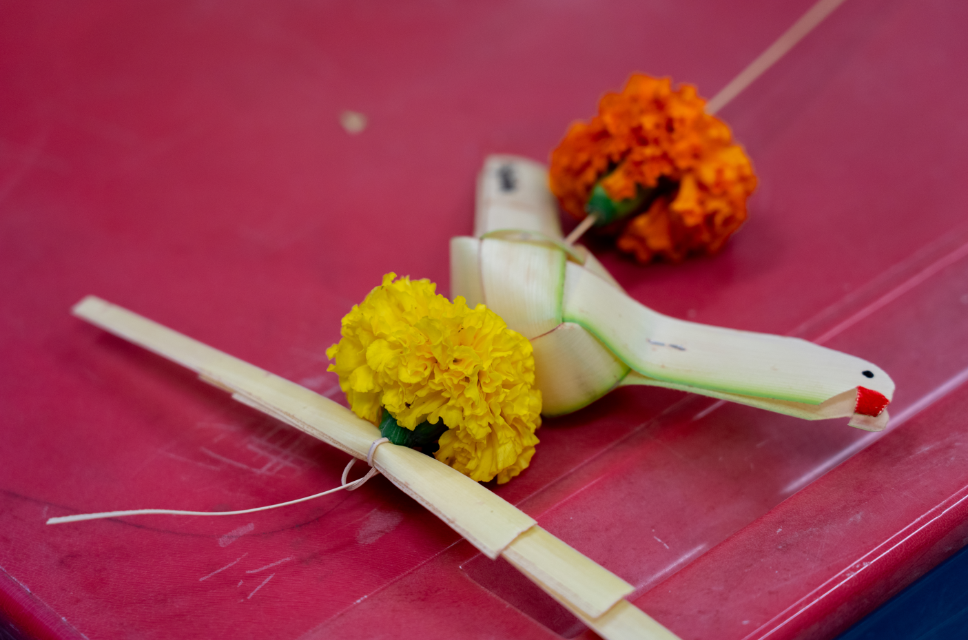 Small woven palm-leaf bird with red beak on pink surface, beside yellow and orange marigolds