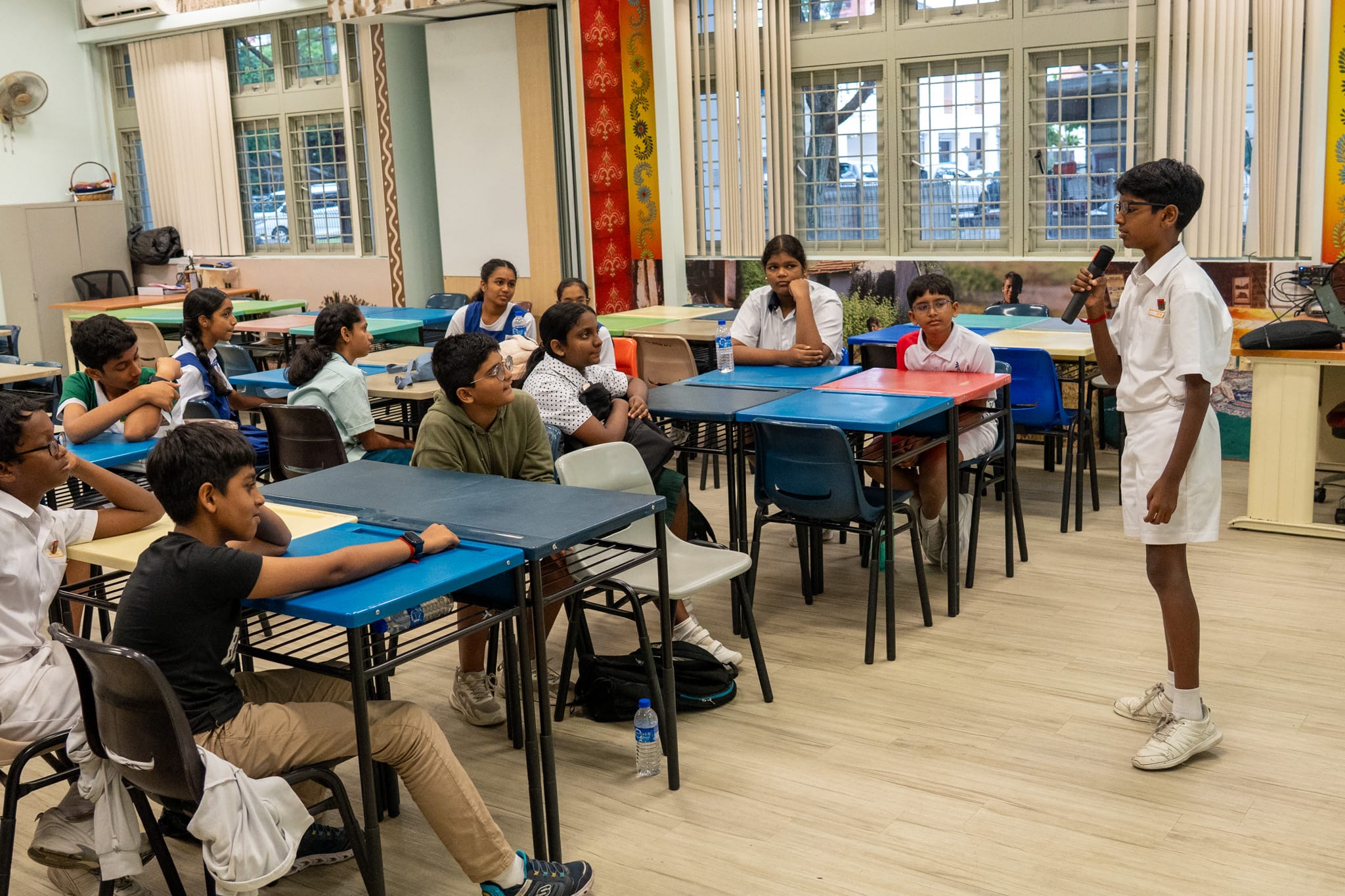 Schoolboy stands with microphone speaking to classmates seated at colorful tables in a classroom.