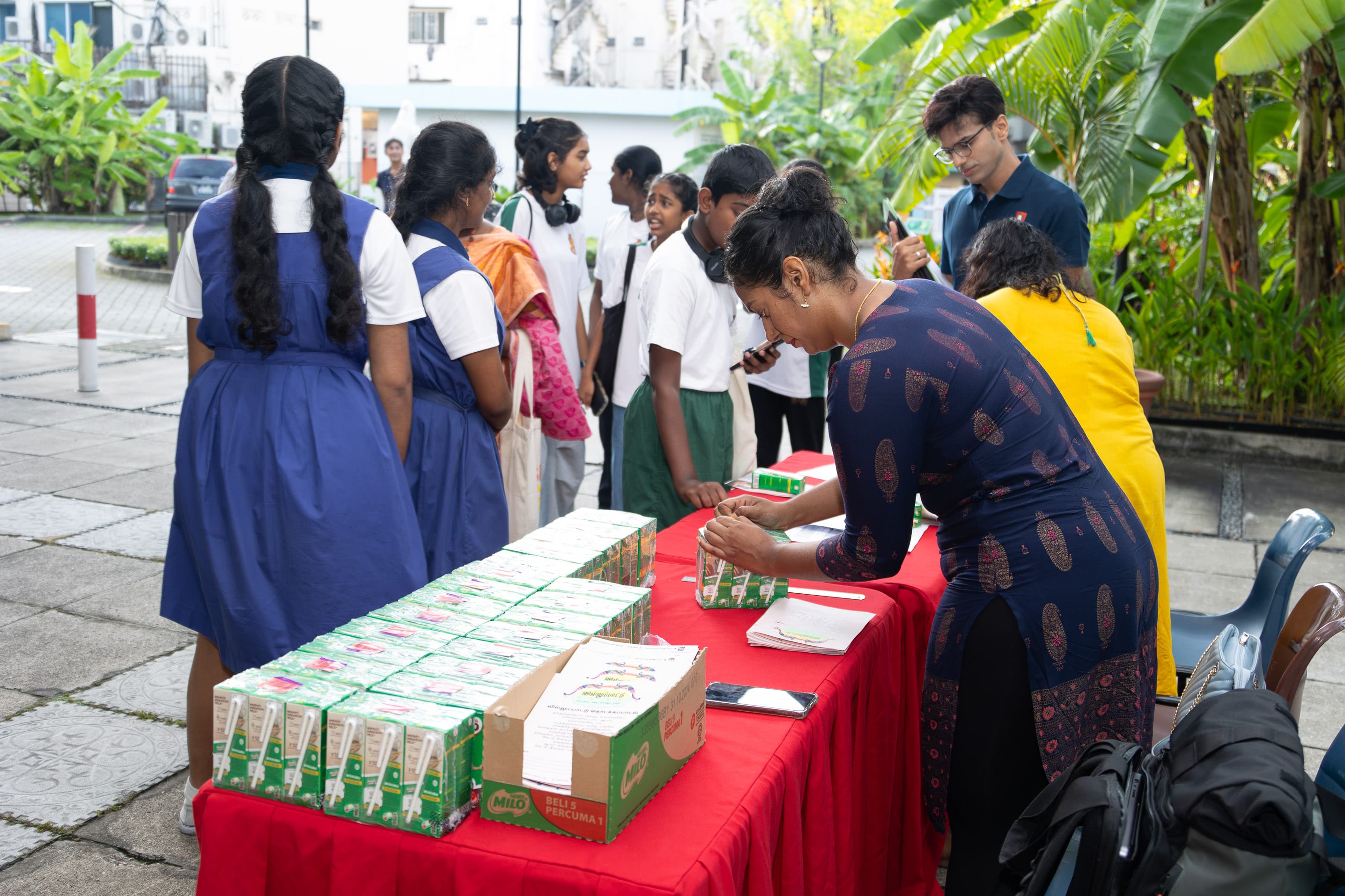 Students line up at an outdoor table with drink cartons as two women prepare items and write on papers.
