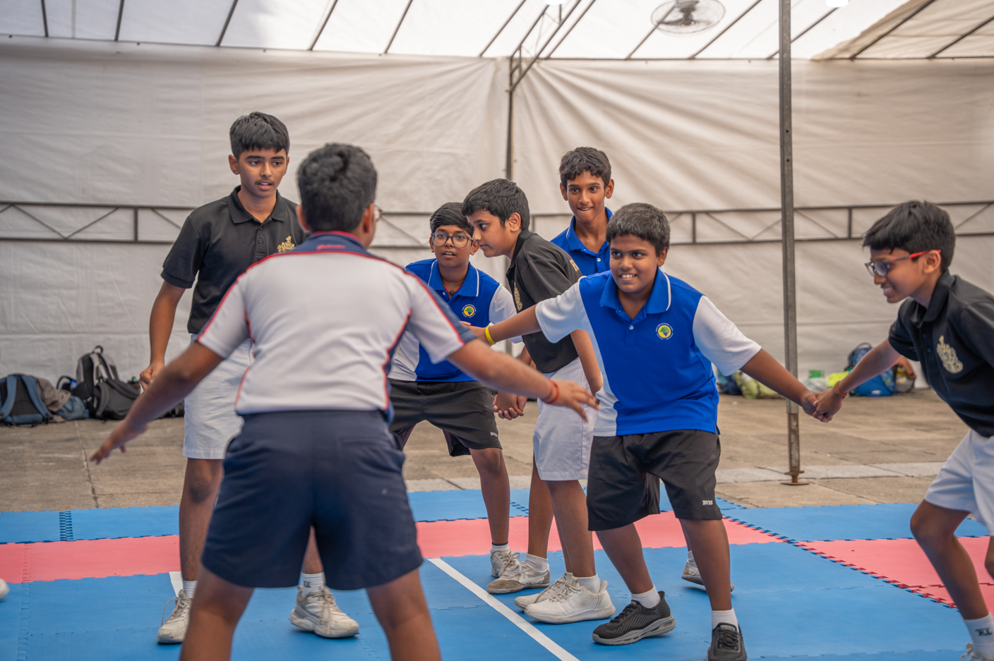 Group of boys on blue mats holding hands and crouching as they play a team game indoors.