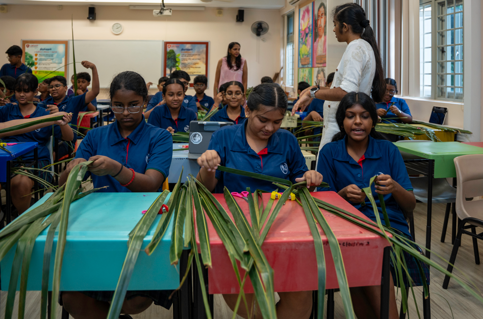 Students in blue uniforms weave long green leaves at desks while a teacher stands nearby in a classroom.