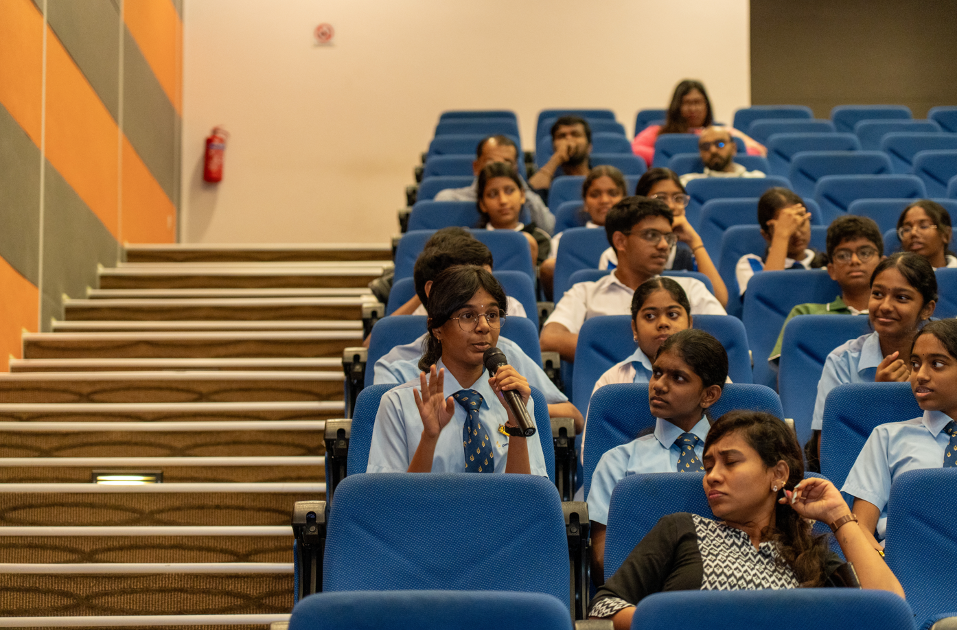 Student in uniform speaks into a microphone in an auditorium with blue seats and classmates listening.